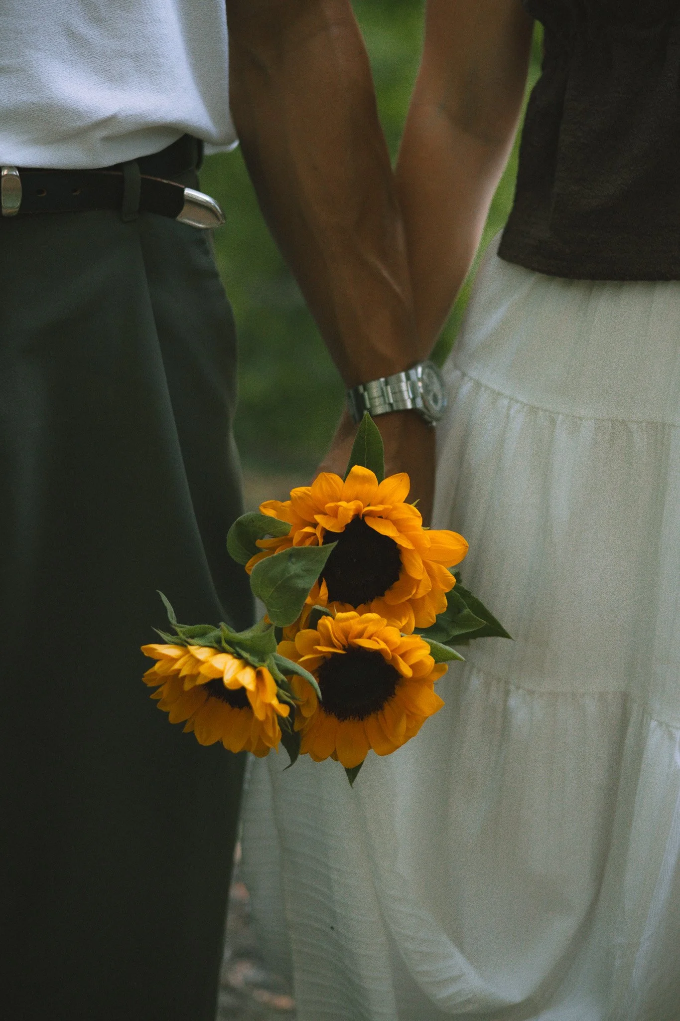 Deux personnes tenant des mains, l'une portant une montre et tenant un bouquet de tournesols. La photo est prise en gros plan, en mettant en évidence leurs bras et mains.