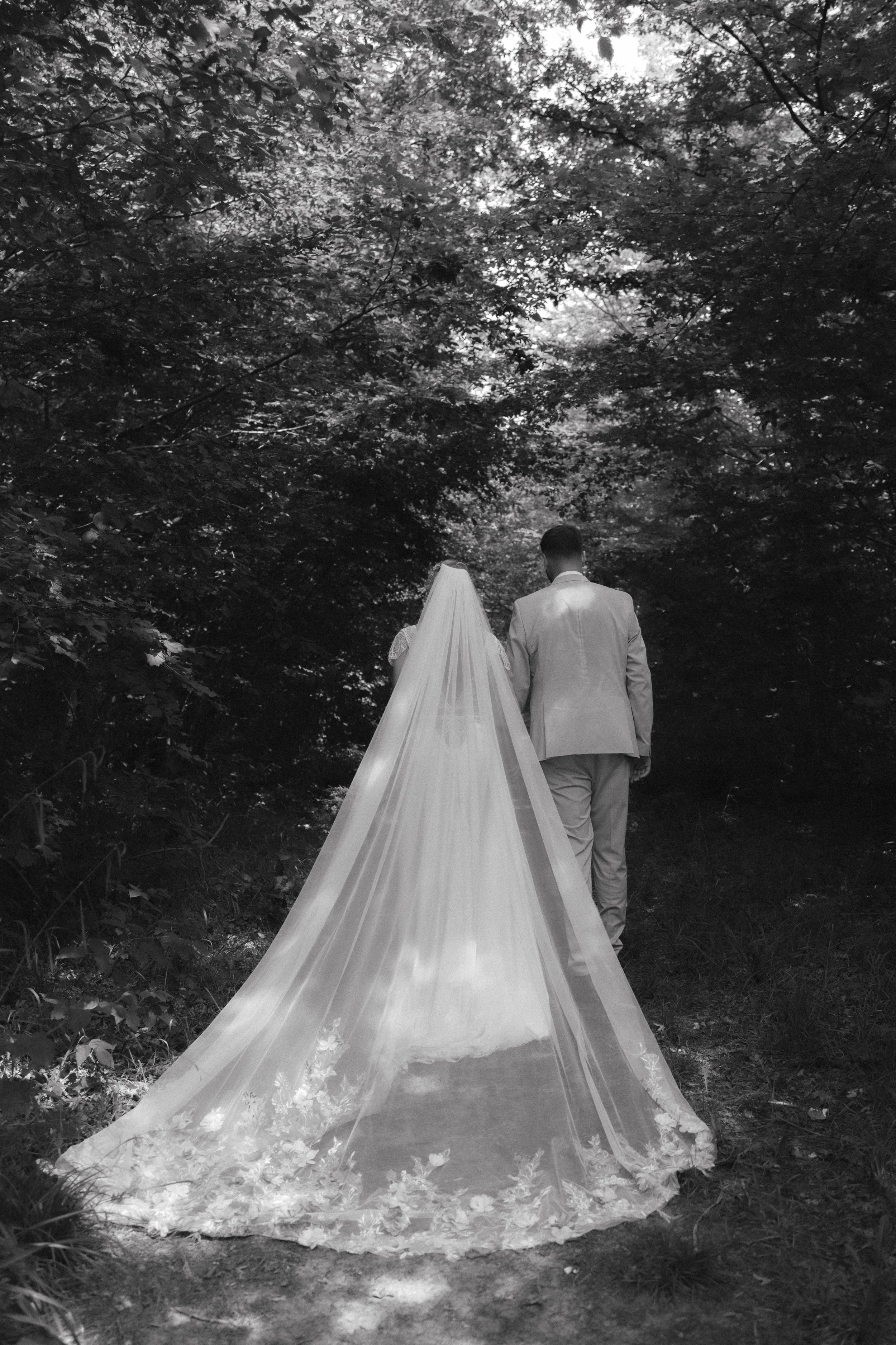 Un couple de mariés marche dans une forêt, vue de dos, la mariée porte une robe de mariée longue et fluide, le marié un costume blanc.
