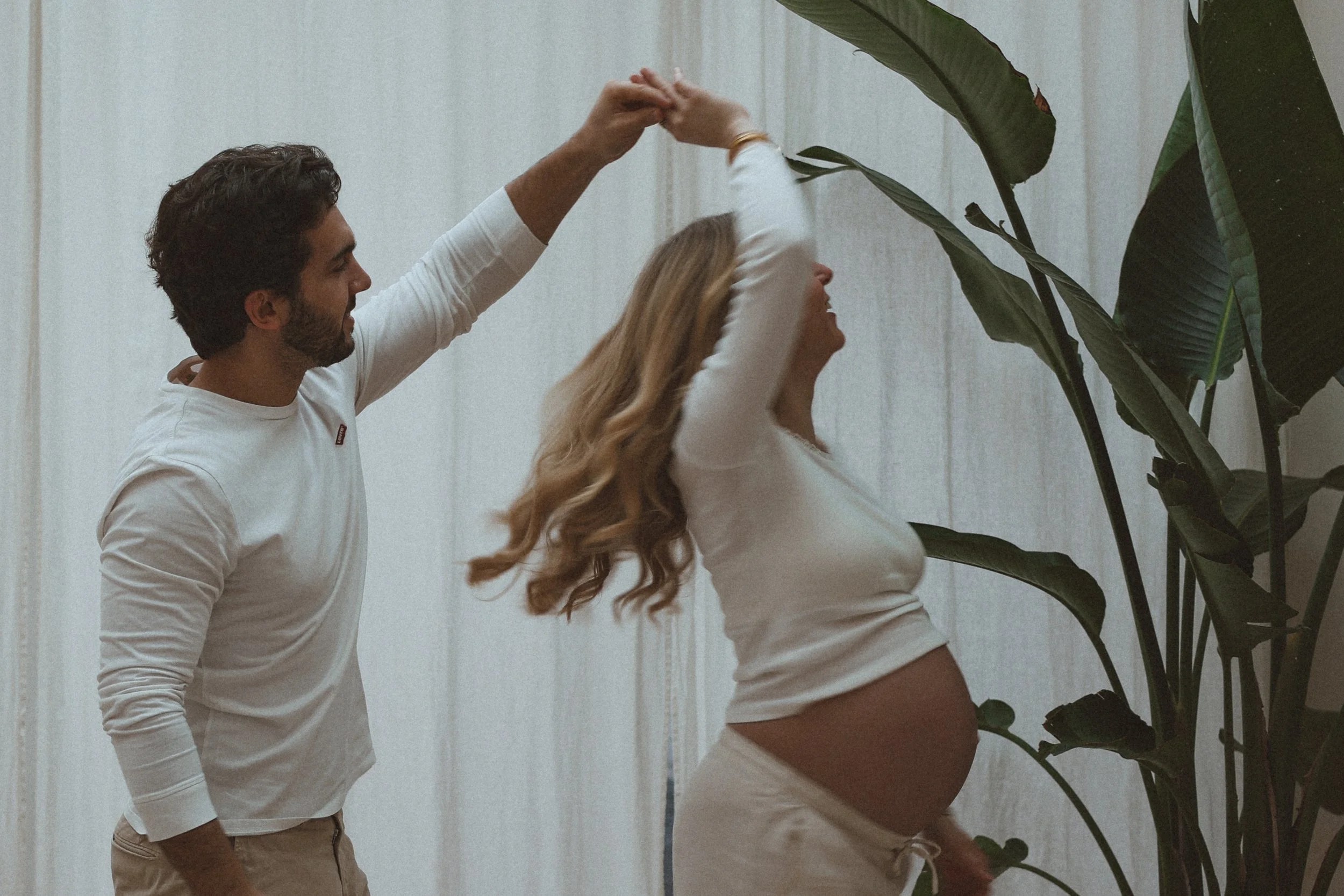Un homme et une femme dansent ensemble, tenant leurs mains levées en haut, dans un intérieur avec un mur blanc et des plantes vertes.