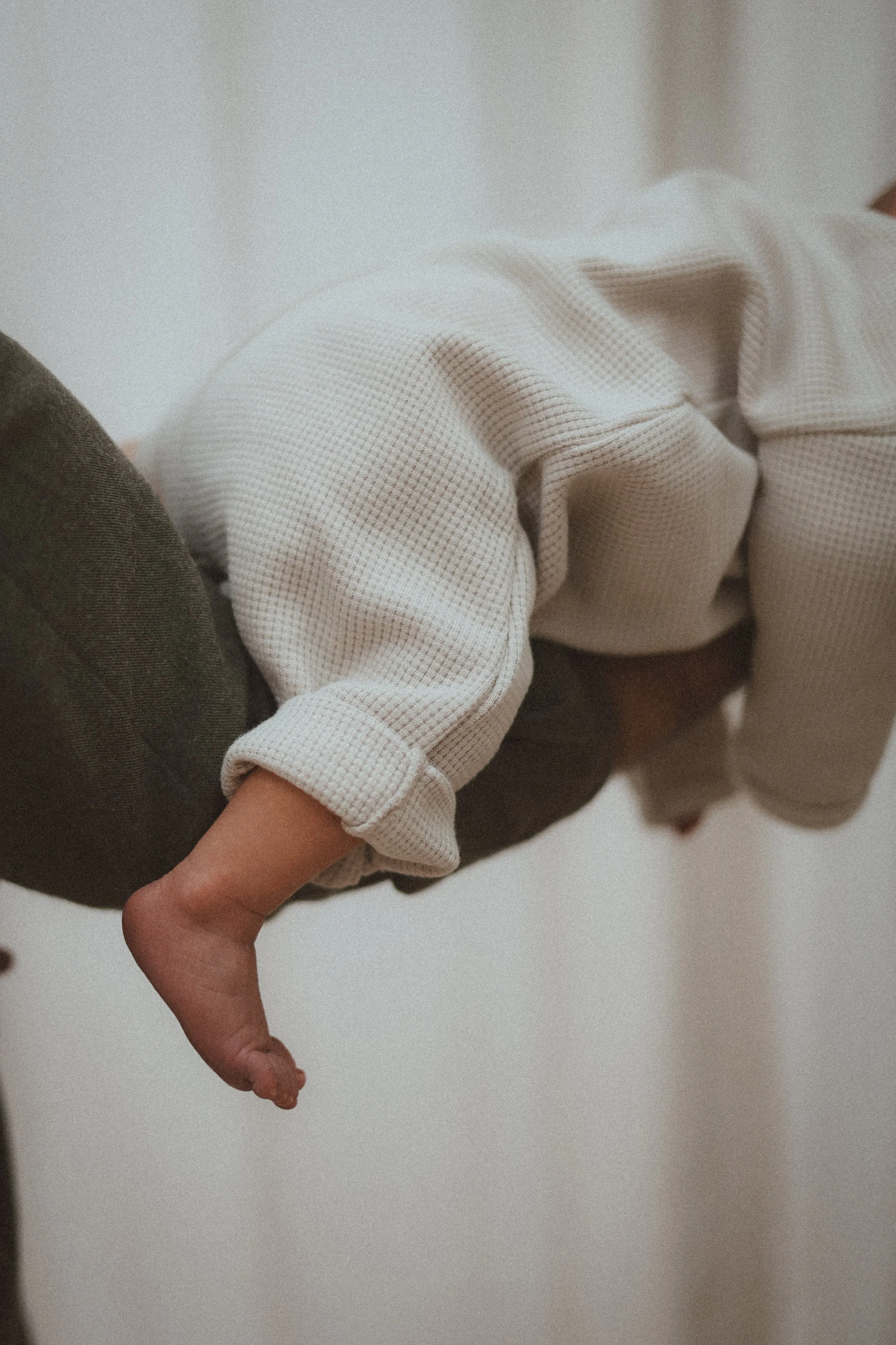 Un bébé suspendu dans les bras d'un adulte, avec un bras visible portant une chemise à manches longues beige à petits carreaux, contre un fond blanc.