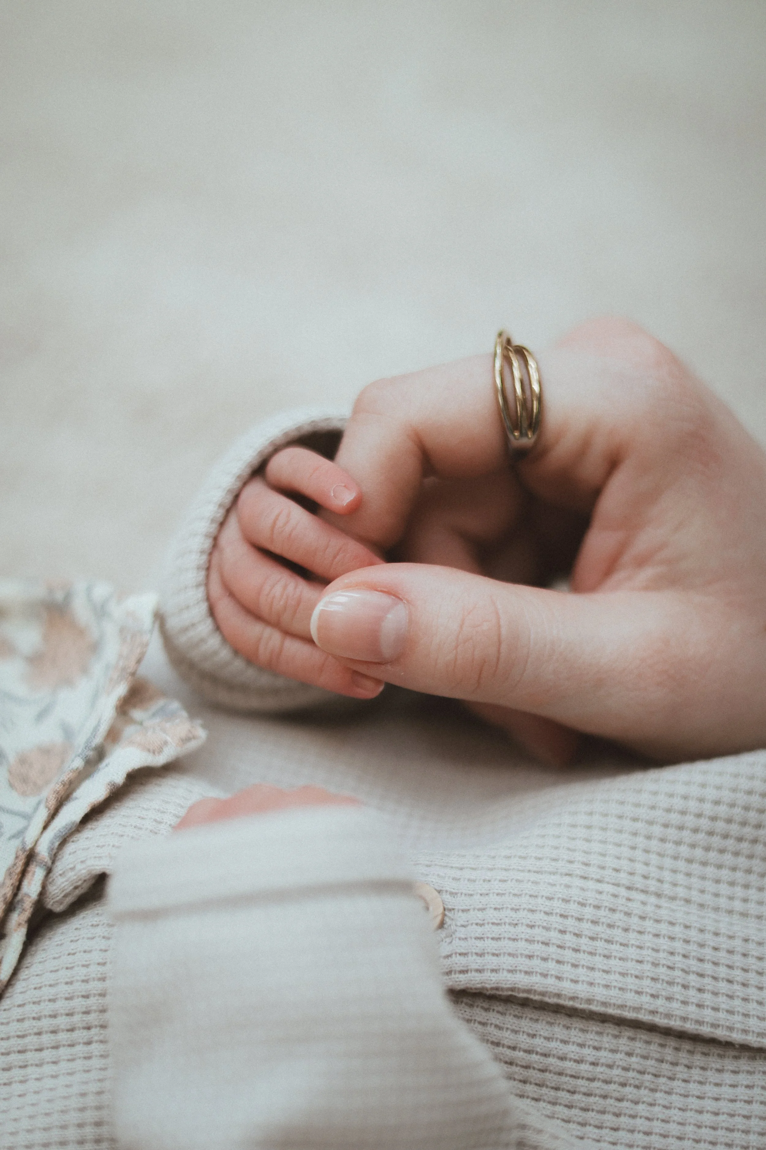 Une main d'adulte avec une bague dorée tient doucement la main d'un bébé. Le bébé porte une tenue en tricot beige avec une couture visible et un ruban en velours rose. Le fond est neutre avec un tissu beige.
