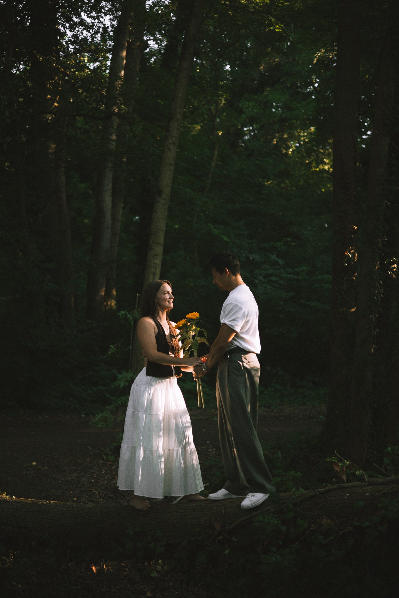 Un couple se tenant la main dans une forêt, l'homme présente un bouquet de fleurs à la femme, tous deux souriants, dans une scène romantique en plein air