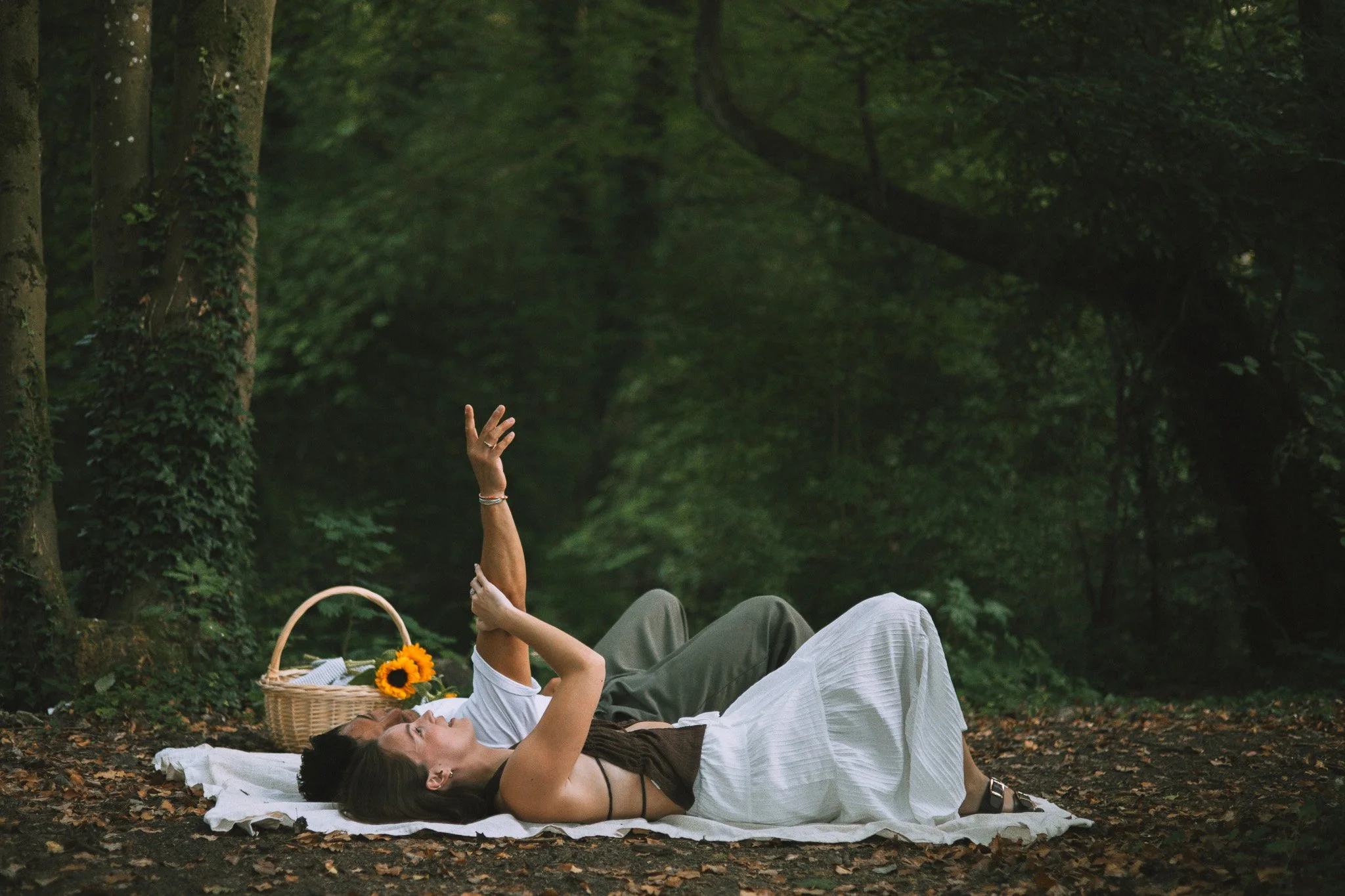 Deux femmes se reposent dans une forêt, allongées sur une couverture blanche, l'une tient une branche ou un objet et l'autre lève la main. Il y a un panier rempli de fleurs de tournesol à côté d'elles.