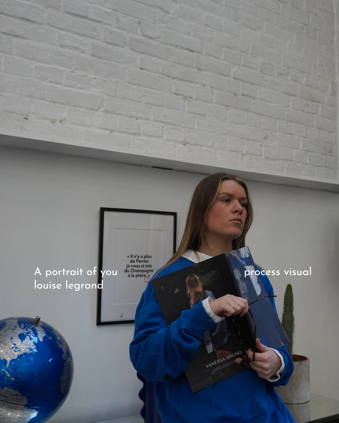 Une jeune femme avec des cheveux longs, portant une veste bleue, tient un magazine ou un livre noir dans ses mains. Elle se trouve dans une pièce moderne avec un mur blanc en briques apparentes au plafond et une plante en pot à l'arrière. Un globe terrestre est visible à gauche.