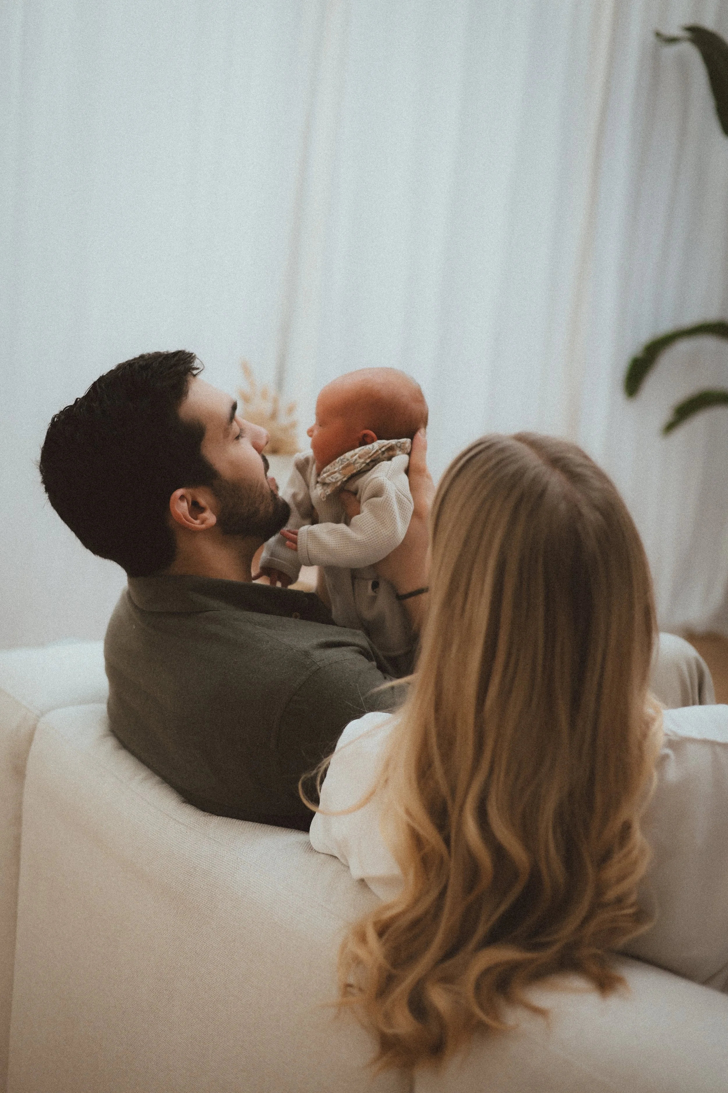 Un homme avec une barbe tient un bébé face à lui. Une femme avec de longs cheveux blonds regarde la scène, assise sur un canapé beige. Le fond comporte des rideaux blancs et une plante verte.