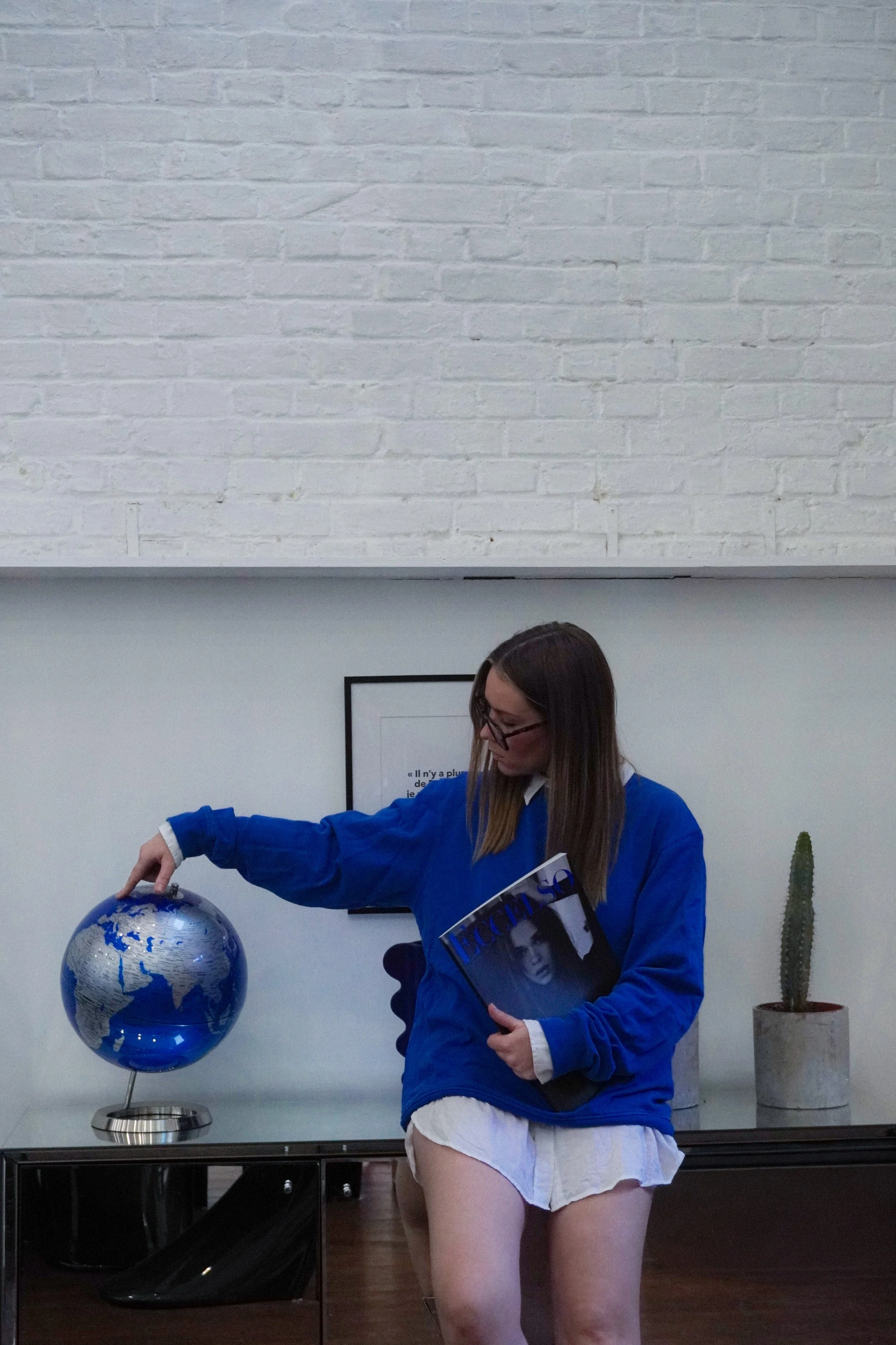 Une jeune femme portant une veste bleue et une jupe courte blanche, tenant un magazine et touchant un globe terrestre.