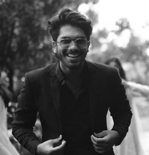 A smiling man wearing glasses and a suit outdoors in a park.