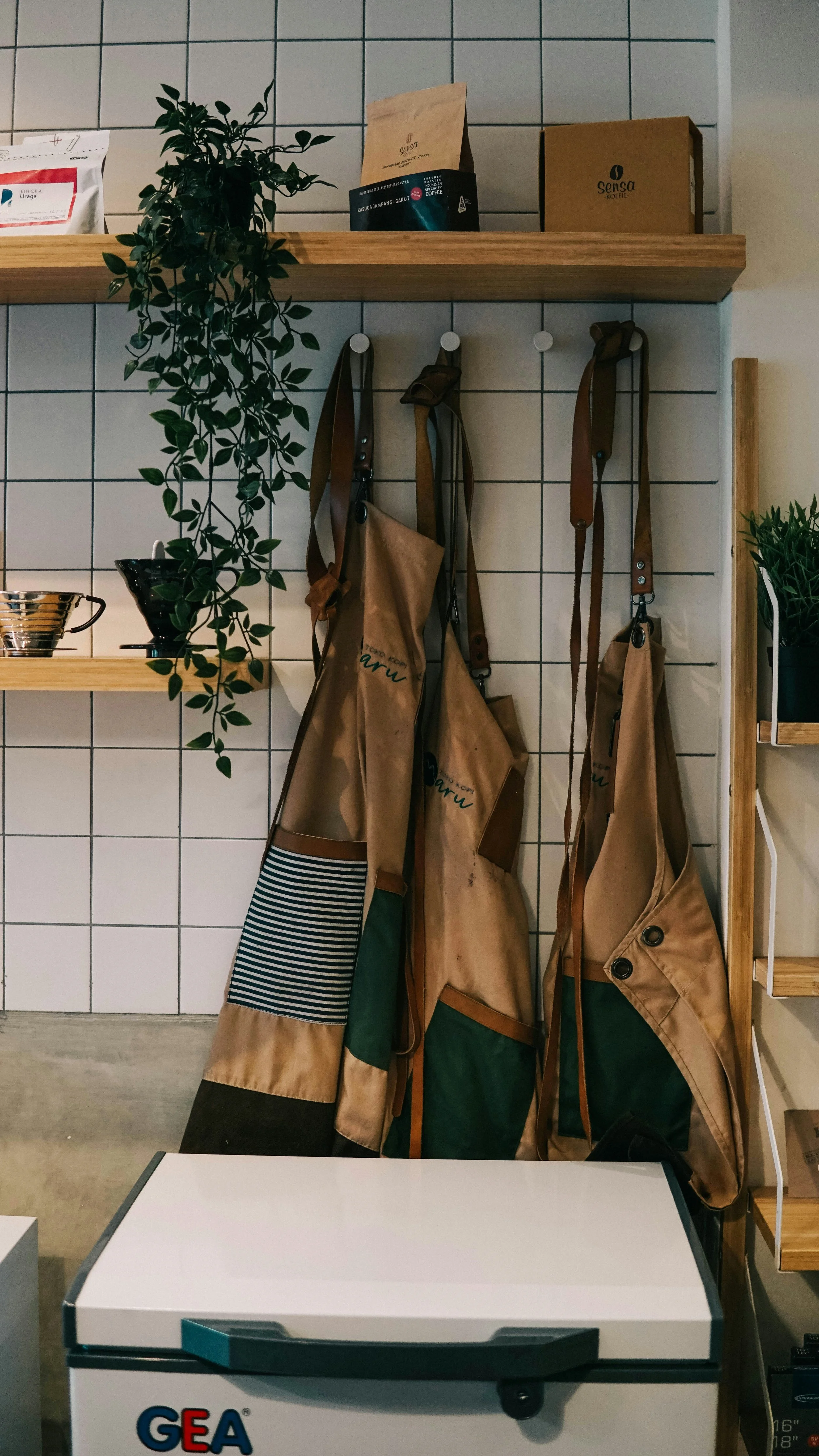 Shelves with hanging aprons, potted plants, coffee bags, and cartons in a cafe or kitchen setting with tiled wall background.
