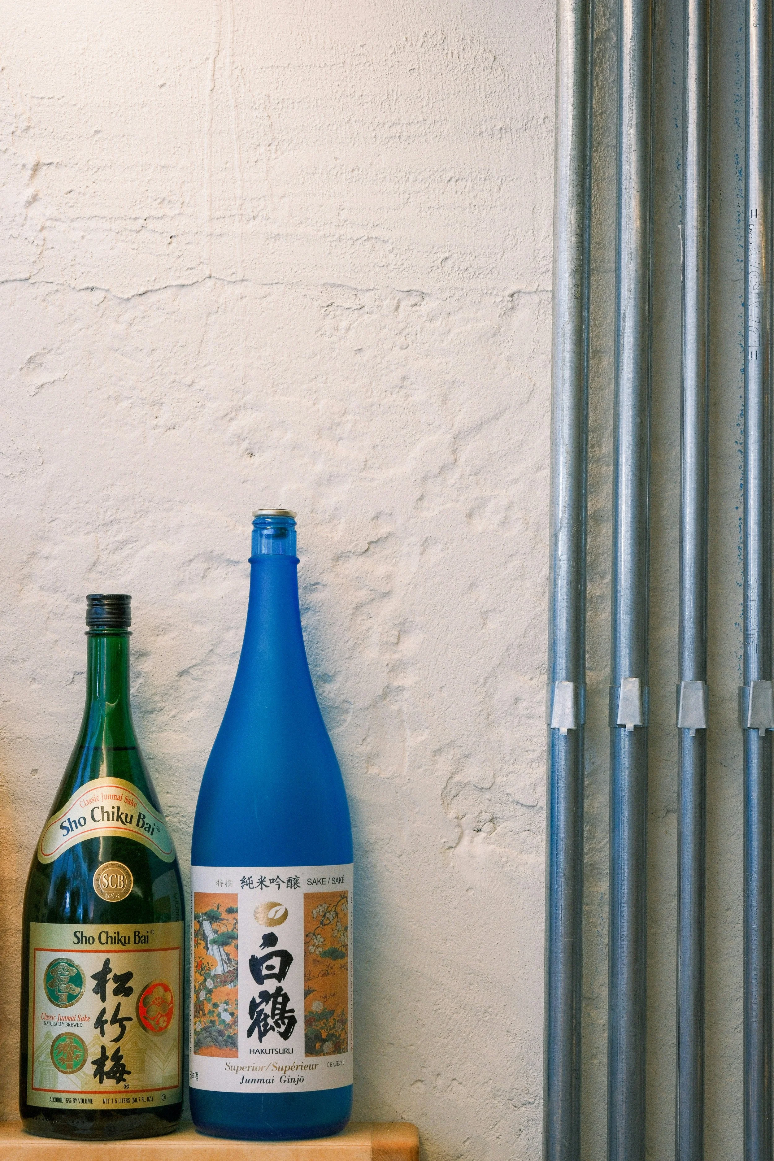 Three bottles of sake and a stack of metal pipes against a beige textured wall.