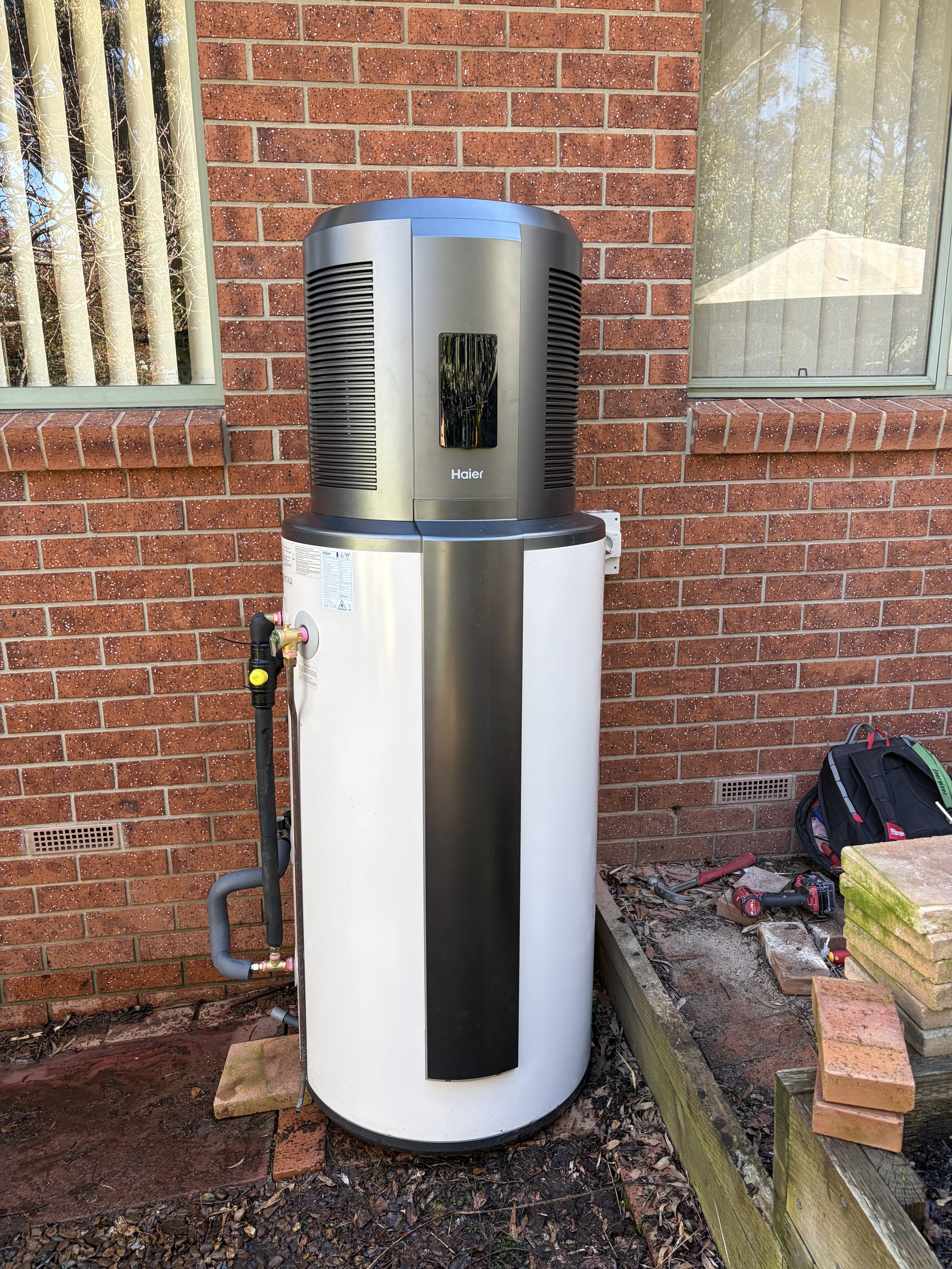 An outdoor scene showing a large, modern water heater heat pump. The water heater has a black and metallic finish with the brand name 'Haier' and is connected to pipes and a faucet.