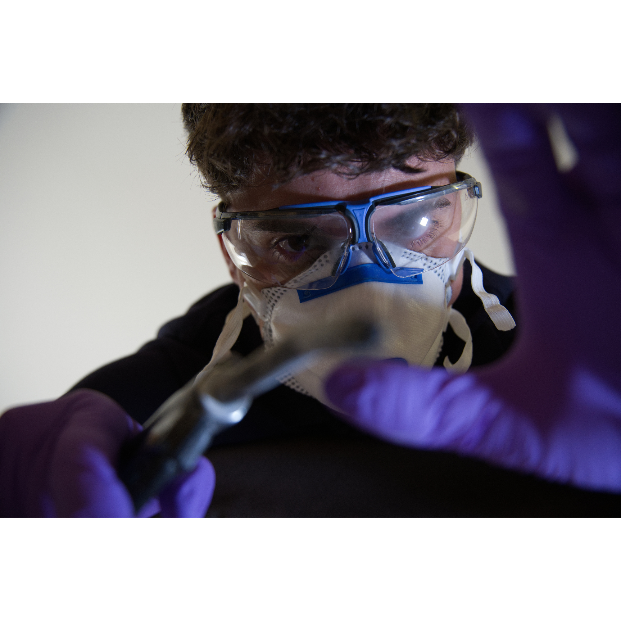 Ein junger Mensch trägt eine Schutzbrille und eine Mundschutzmaske, scheint eine technische oder wissenschaftliche Tätigkeit auszuführen.