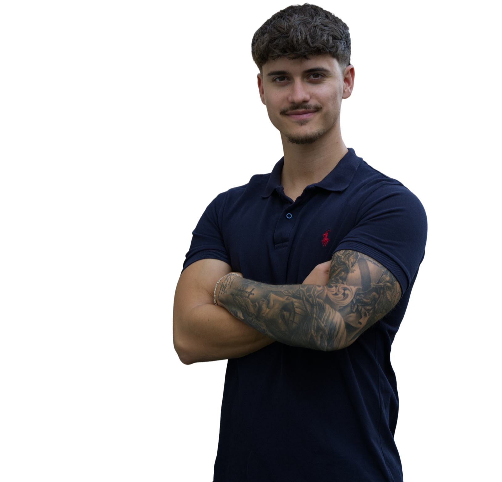 Ein junger Mann mit kurzen, lockigen braunen Haaren und einem leichten Bart, der eine dunkle Poloshirt trägt, versetzt die Arme vor der Brust und zeigt ein Lächeln. Sein linker Arm ist mit detaillierten Tattoos bedeckt.