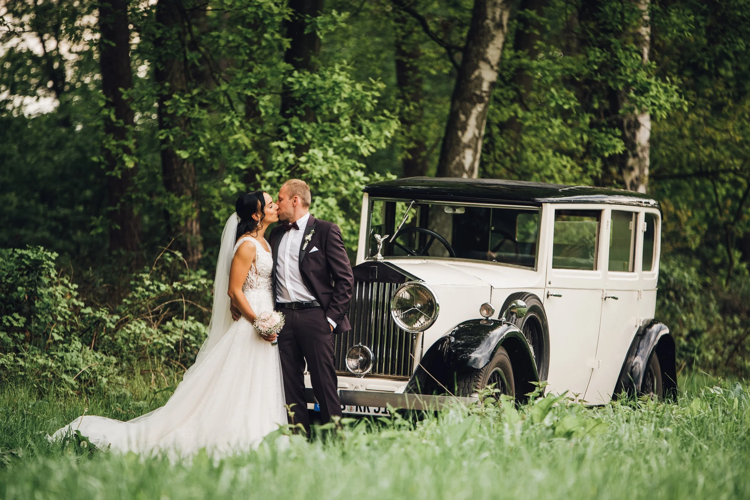 Ein Hochzeitspaar küss sich vor einem alten weißen Auto in einem Wald.