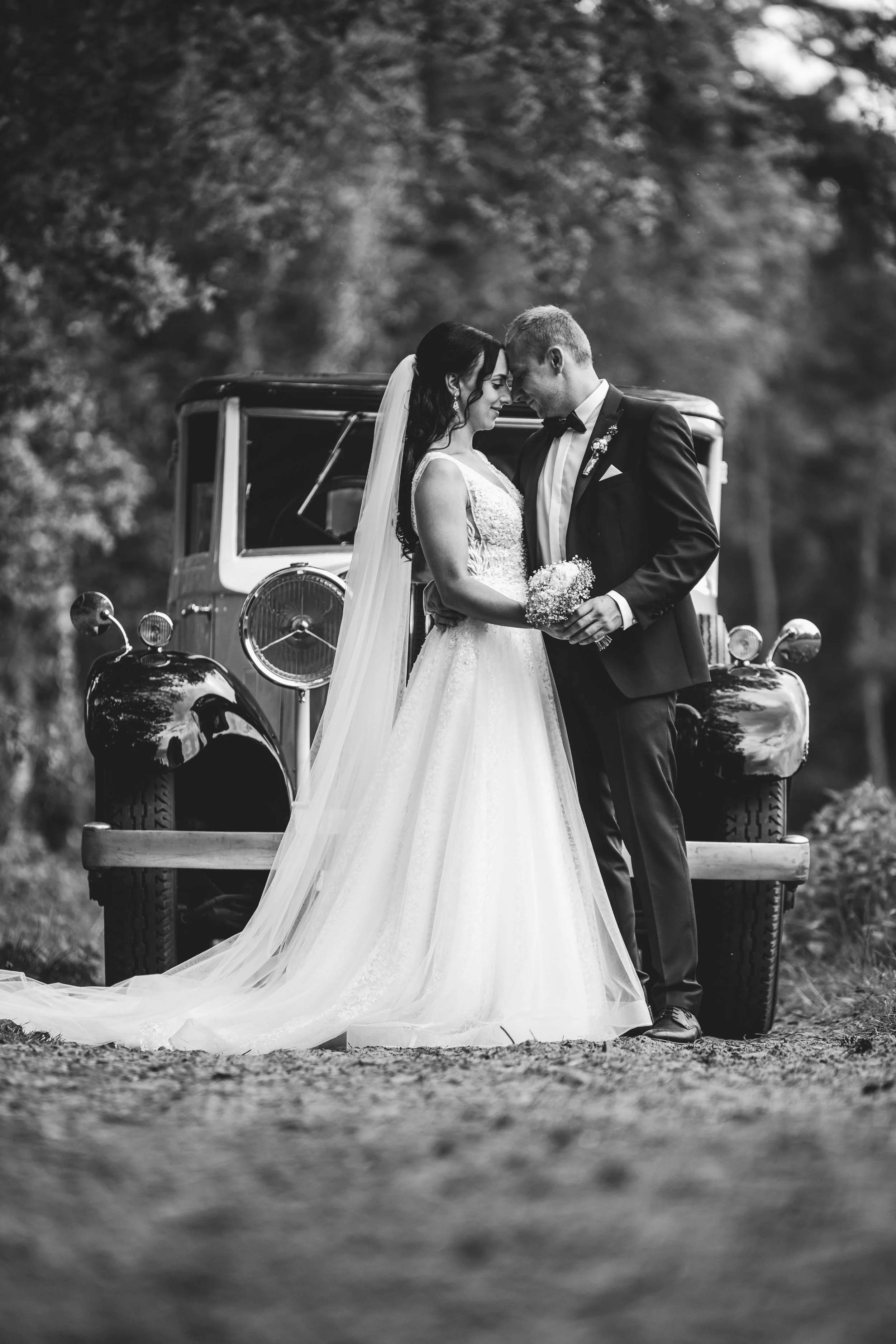 Ein Brautpaar steht sich vor einem alten Auto gegenüber, mit den Stirn aneinander, bei einer Hochzeit im Freien, schwarz-weiß Fotografie.