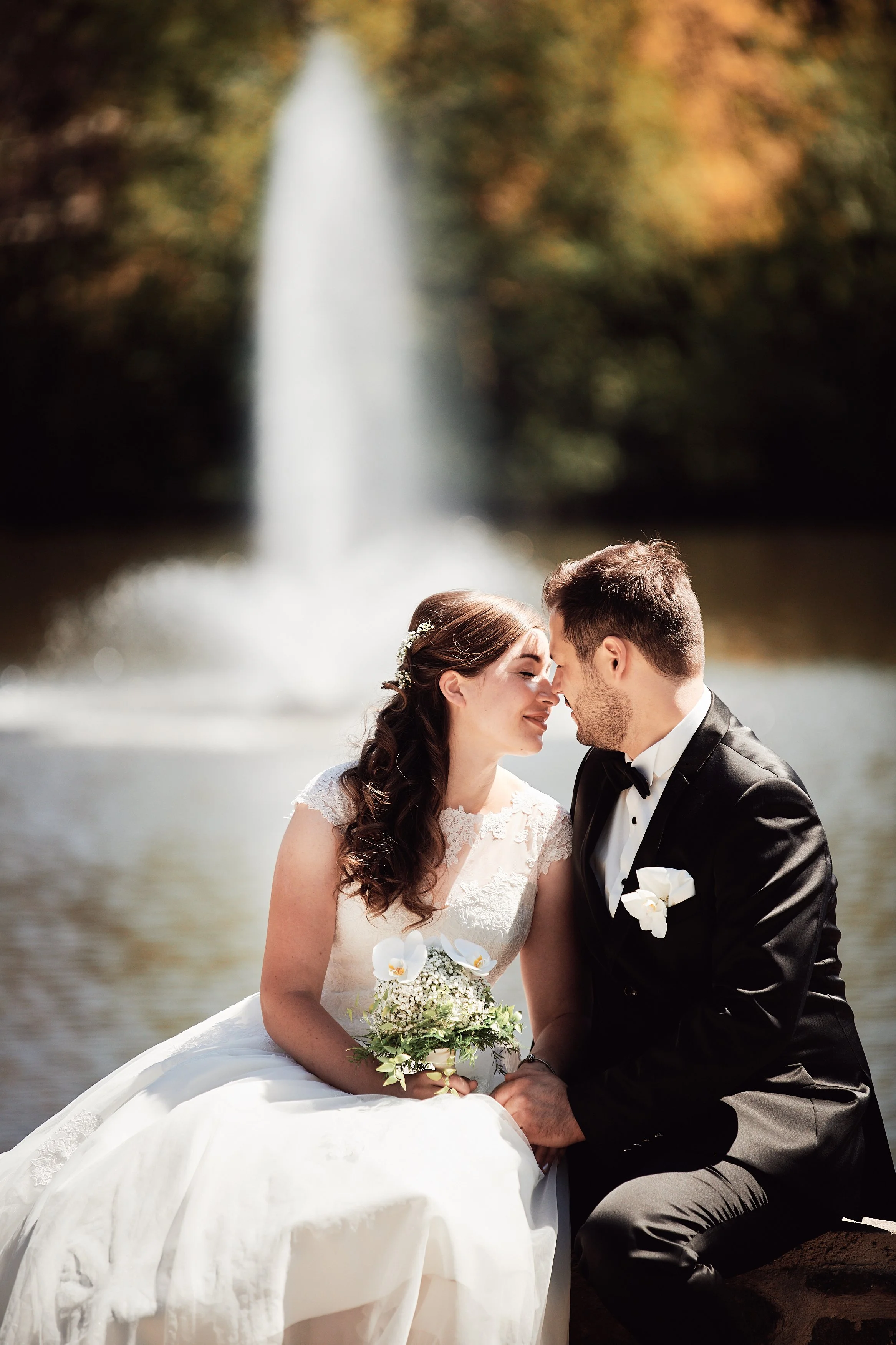 Ein brautpaar sitzt an einem Fluss mit Wasserfall im Hintergrund. Die Frau trägt ein weißes Hochzeitskleid und hält einen Blumenstrauß, der Mann trägt einen schwarzen Anzug mit Fliege. Sie berühren sich sanft an den Stirnen, haben ihre Augen geschlossen und lächeln.