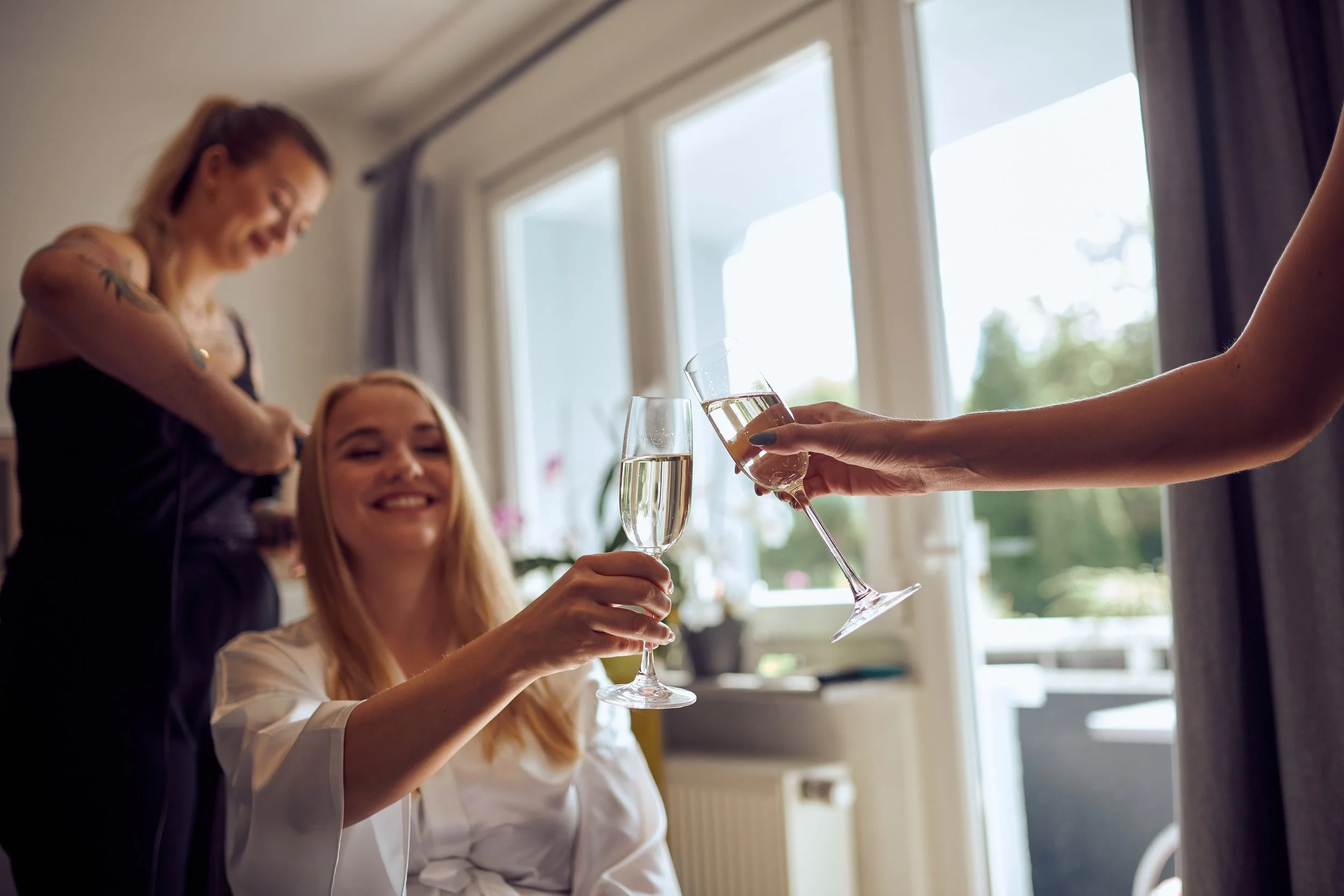 Zwei Frauen stoßen mit Sektgläsern an, eine lächelt, während die andere im Hintergrund steht, vor großen Fenstern mit Vorhängen.