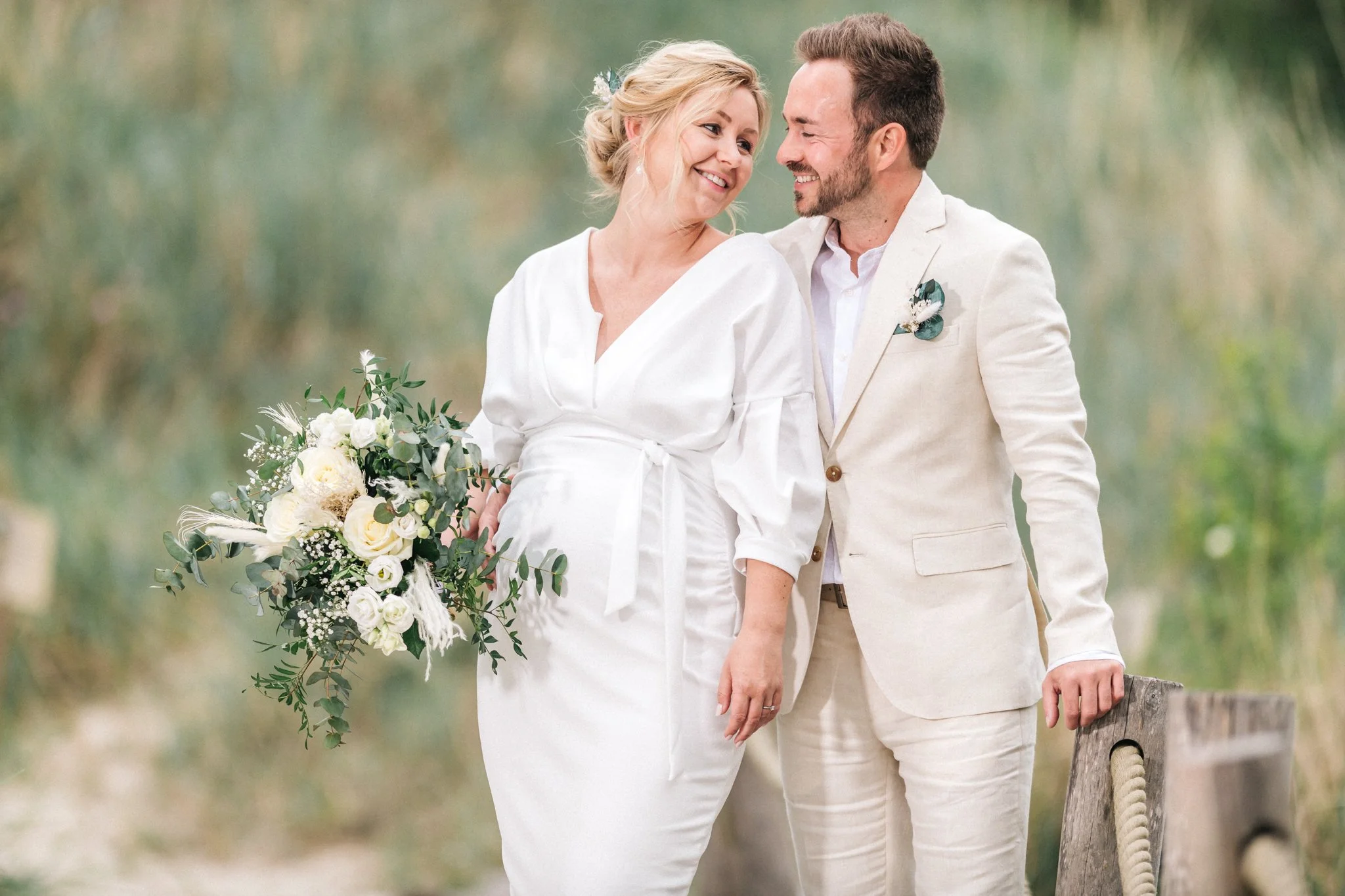 Ein älteres Ehepaar bei einer Hochzeit im Grünen, die Frau trägt ein weißes Kleid, hält einen Blumenstrauß, der Mann trägt einen hellen Anzug und hält eine Anker.