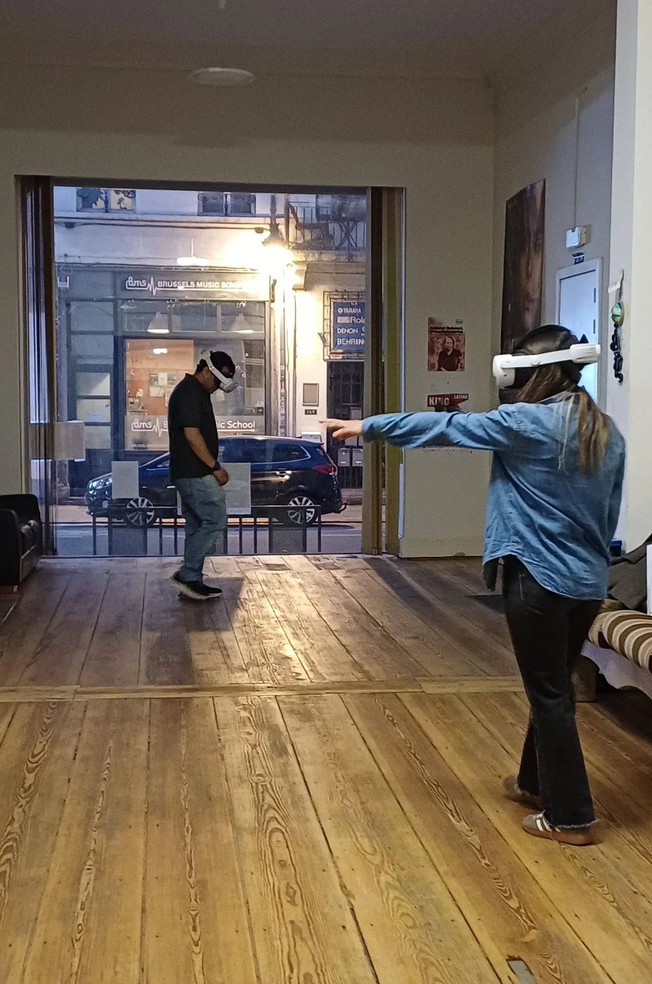 Deux personnes portant des casques de réalité virtuelle dans un espace intérieur avec un grand espace en bois au sol. Une personne montre une direction avec la main, tandis que l'autre est debout près de la fenêtre.