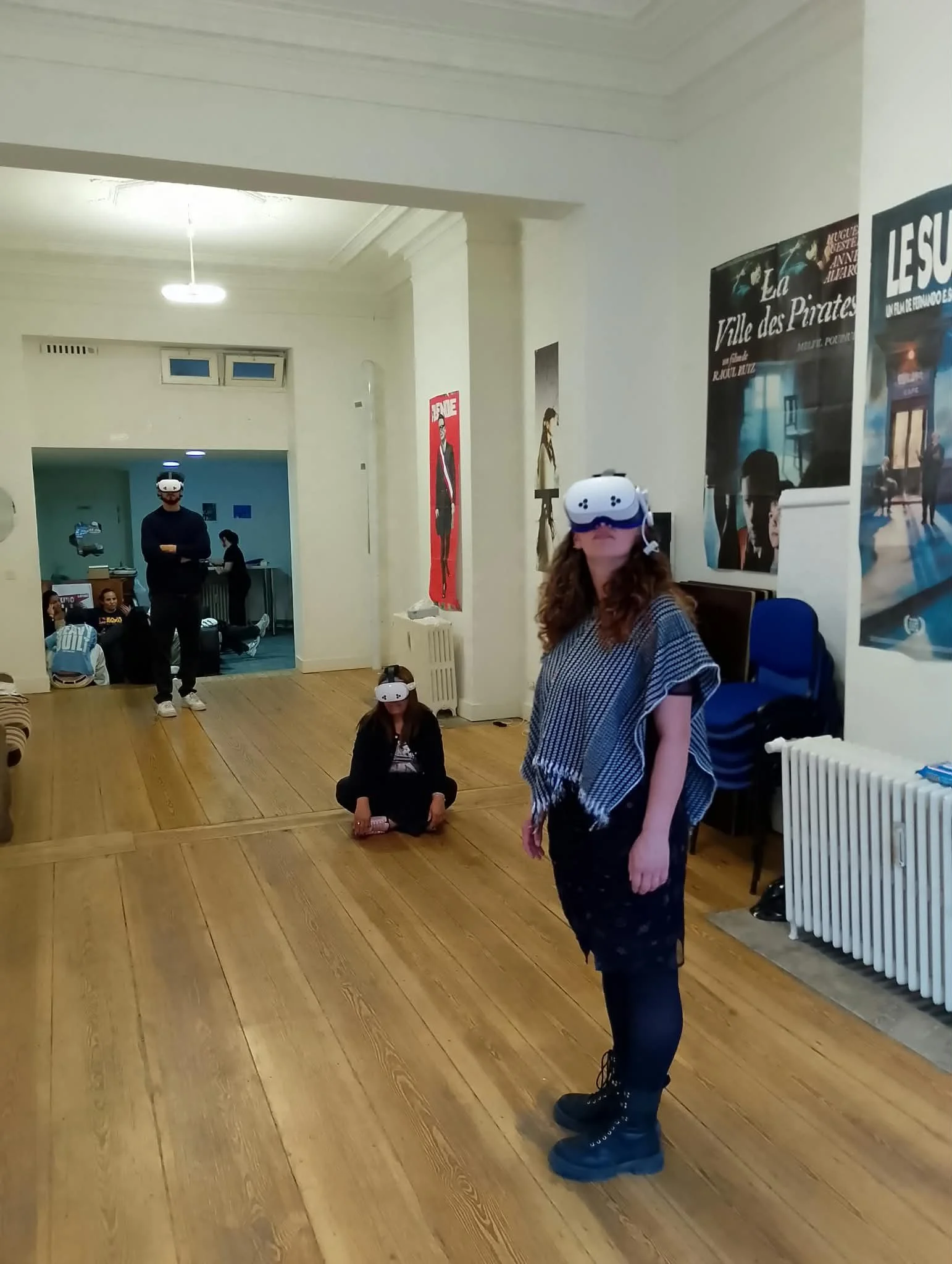 Une femme porte un casque de réalité virtuelle dans une pièce avec d'autres personnes aussi équipées de casquesVR. Certaines personnes sont assises ou debout, d'autres regardent ou participent à une activité immersive.
