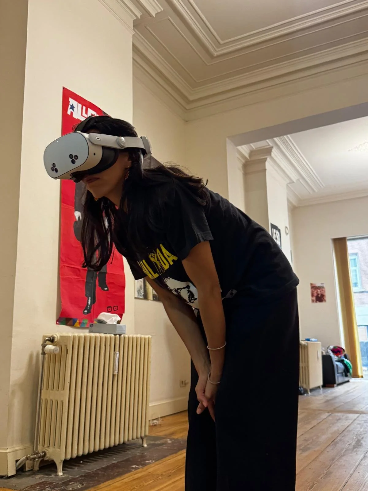 Une femme portant un casque de réalité virtuelle inclinée en avant dans une pièce avec des murs clairs, un radiateur ancien, et une bannière rouge avec un personnage noir et blanc.