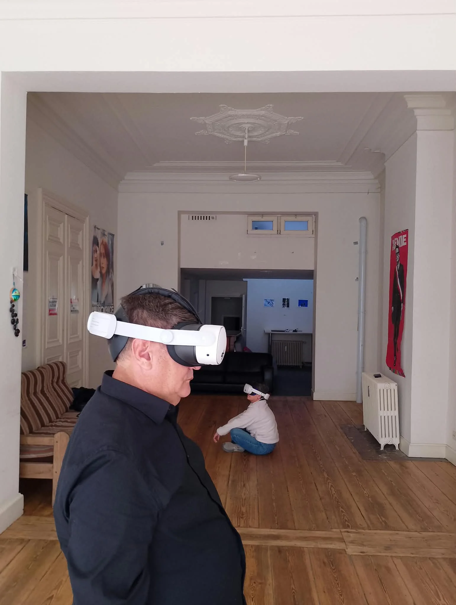 Un homme portant un casque de réalité virtuelle dans une pièce intérieur, avec deux personnes assises sur le sol en arrière-plan.
