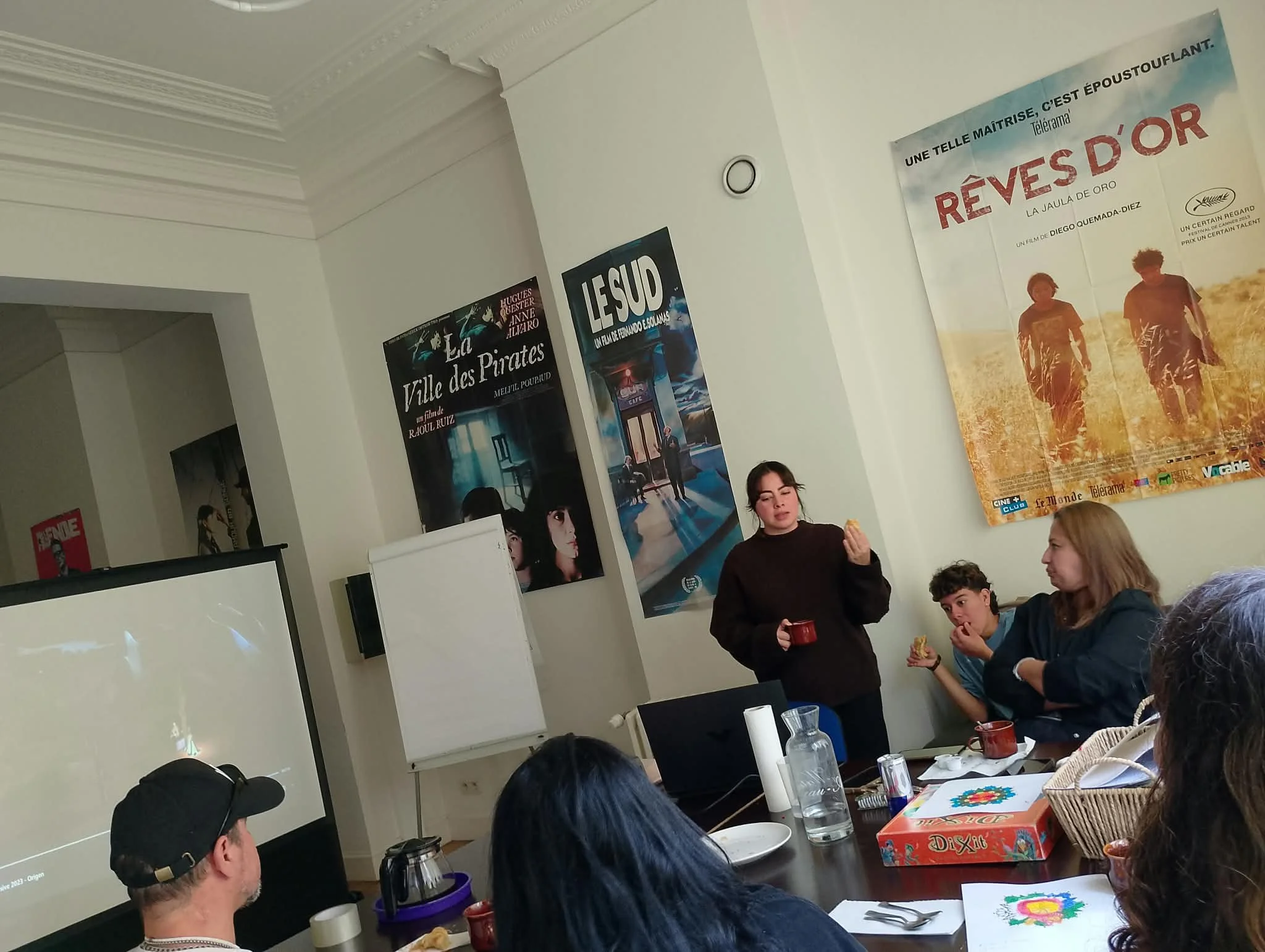 Une salle de réunion avec des personnes assises autour d'une table, regardant un écran. Il y a plusieurs affiches de films sur le mur, dont 'Rêves d'or' et 'Les U.Sud'.