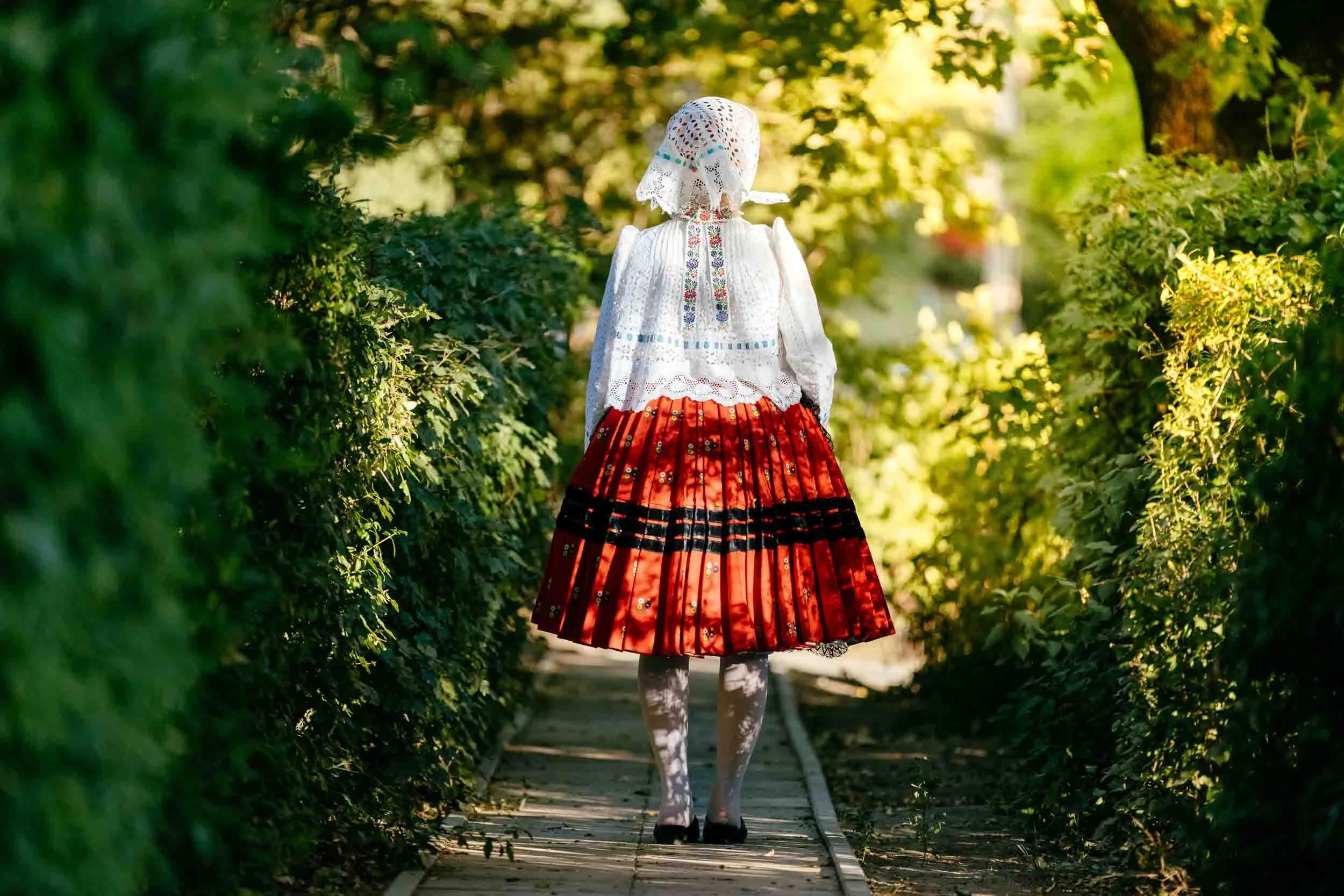 A girl in traditional folk clothing, with a white embroidered blouse and red skirt, walks along a narrow wooden pathway surrounded by lush green foliage, with sunlight filtering through the trees.