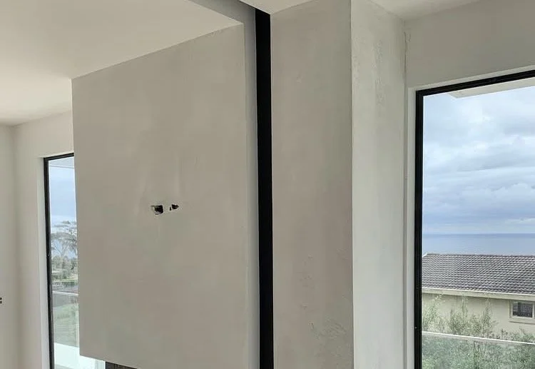 Construction or renovation with unfinished wall, exposed outlets, and large windows showing a cloudy sky and neighboring houses.