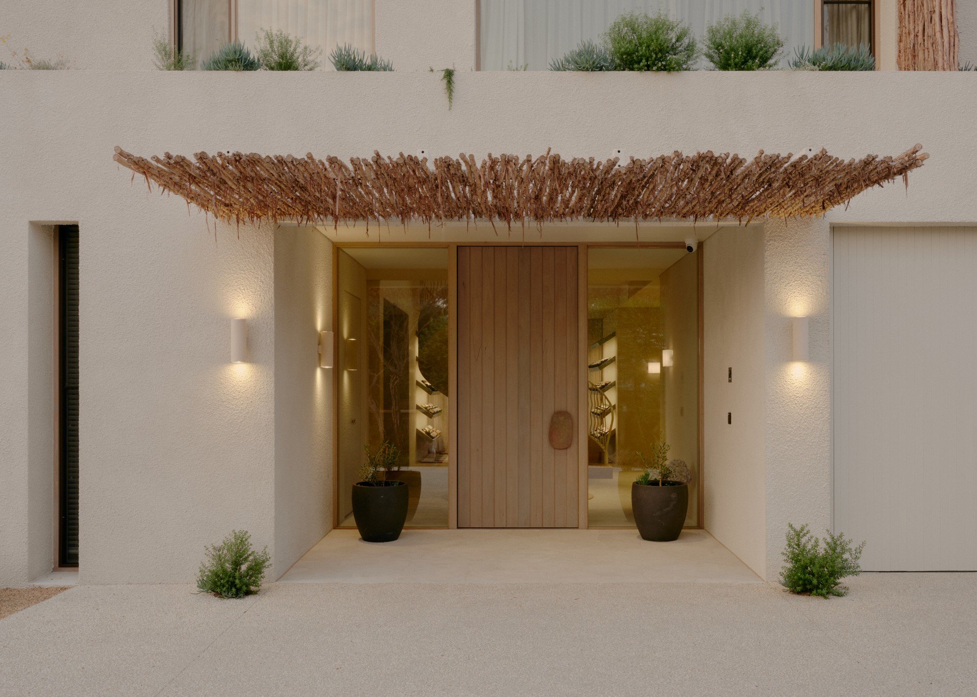 Modern home entrance with beige textured walls, wooden double doors, two large black pots with plants, and small green shrubs on either side. Lights illuminate the entrance.