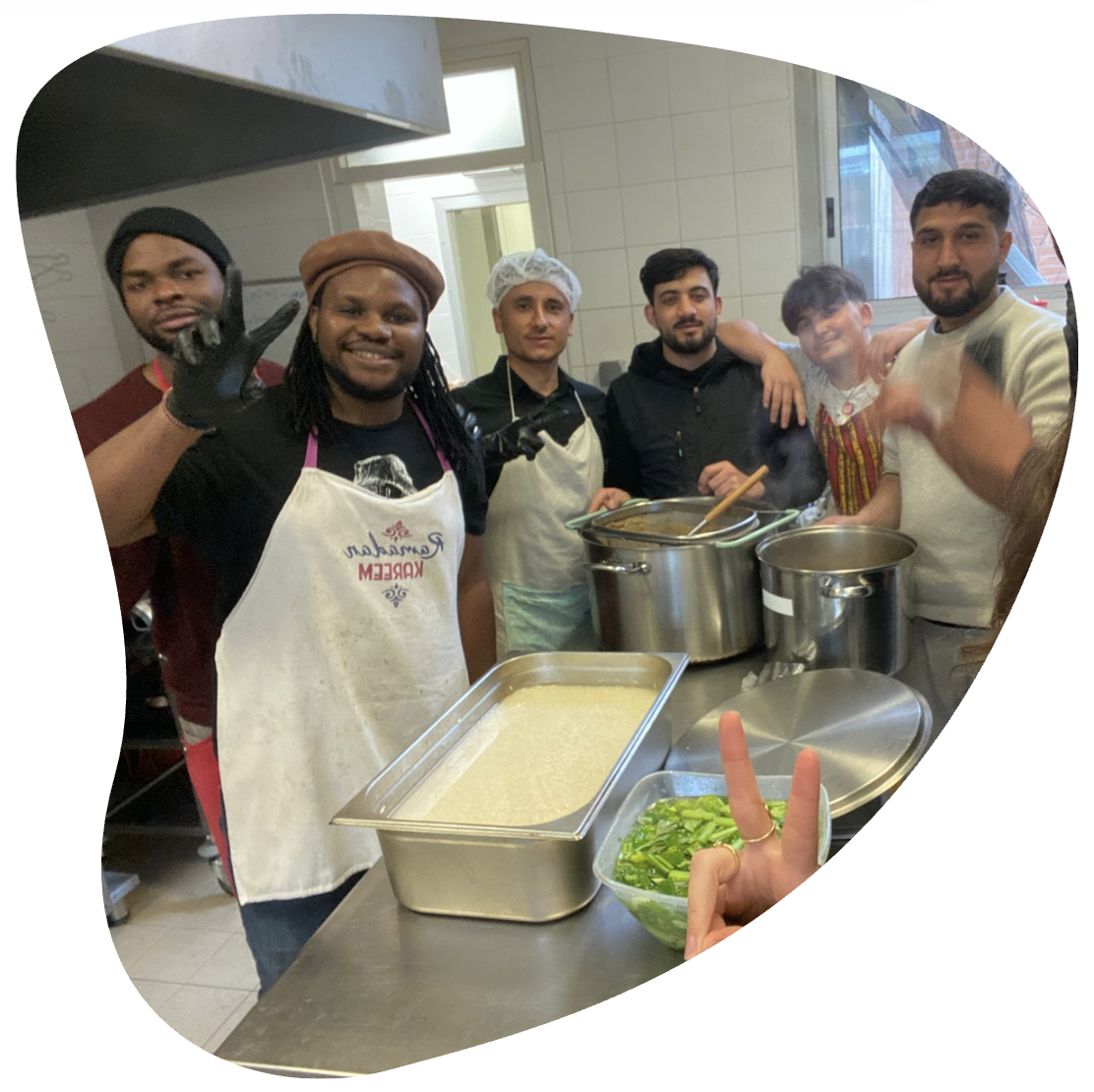 Groupe de sept personnes en cuisine, certains portant des tabliers, posant devant des casseroles et des contenants de nourriture, souriant et faisant des signes de paix ou des gestes amicaux.