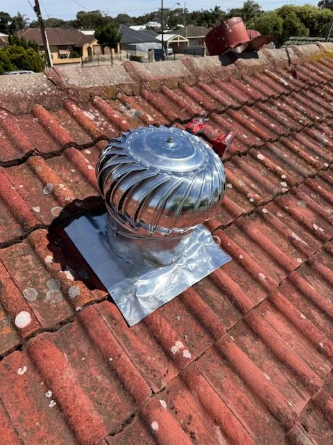 A roof with red tiles featuring a shiny, metallic turbine vent and a red vent cap, with neighboring houses and trees in the background.