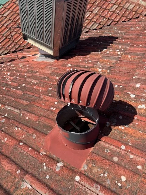 A roof vent with a rotating cover is open on a rooftop with reddish-brown shingles; an air conditioning unit is visible in the background.