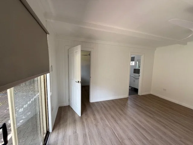 Empty living room with white walls and light wood flooring, sliding glass door with window shade, open door to another room, and a partial view of a bathroom.