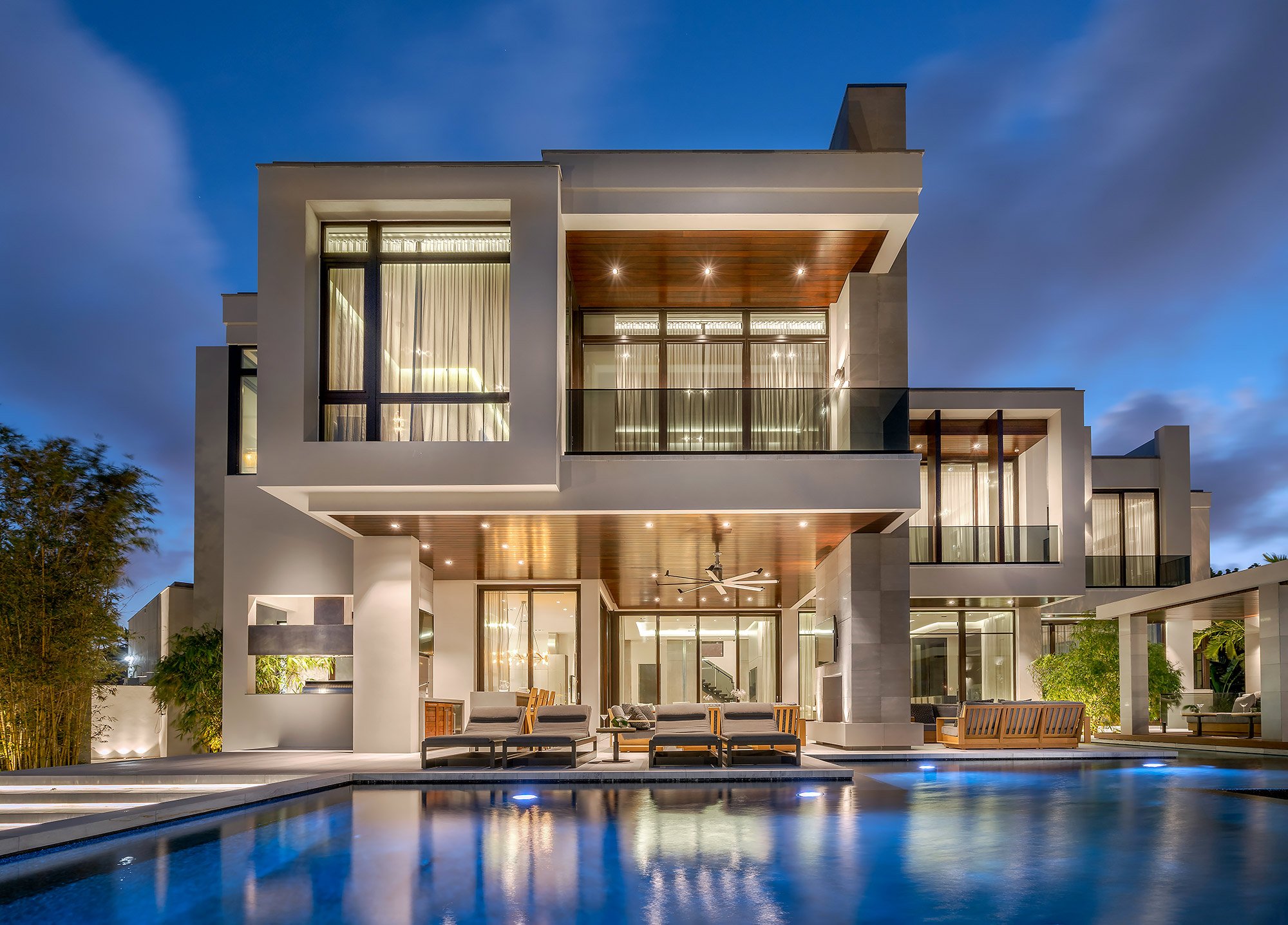 Modern multi-story house with large glass windows and outdoor patio area, illuminated at night, with swimming pool in the foreground, surrounded by trees.