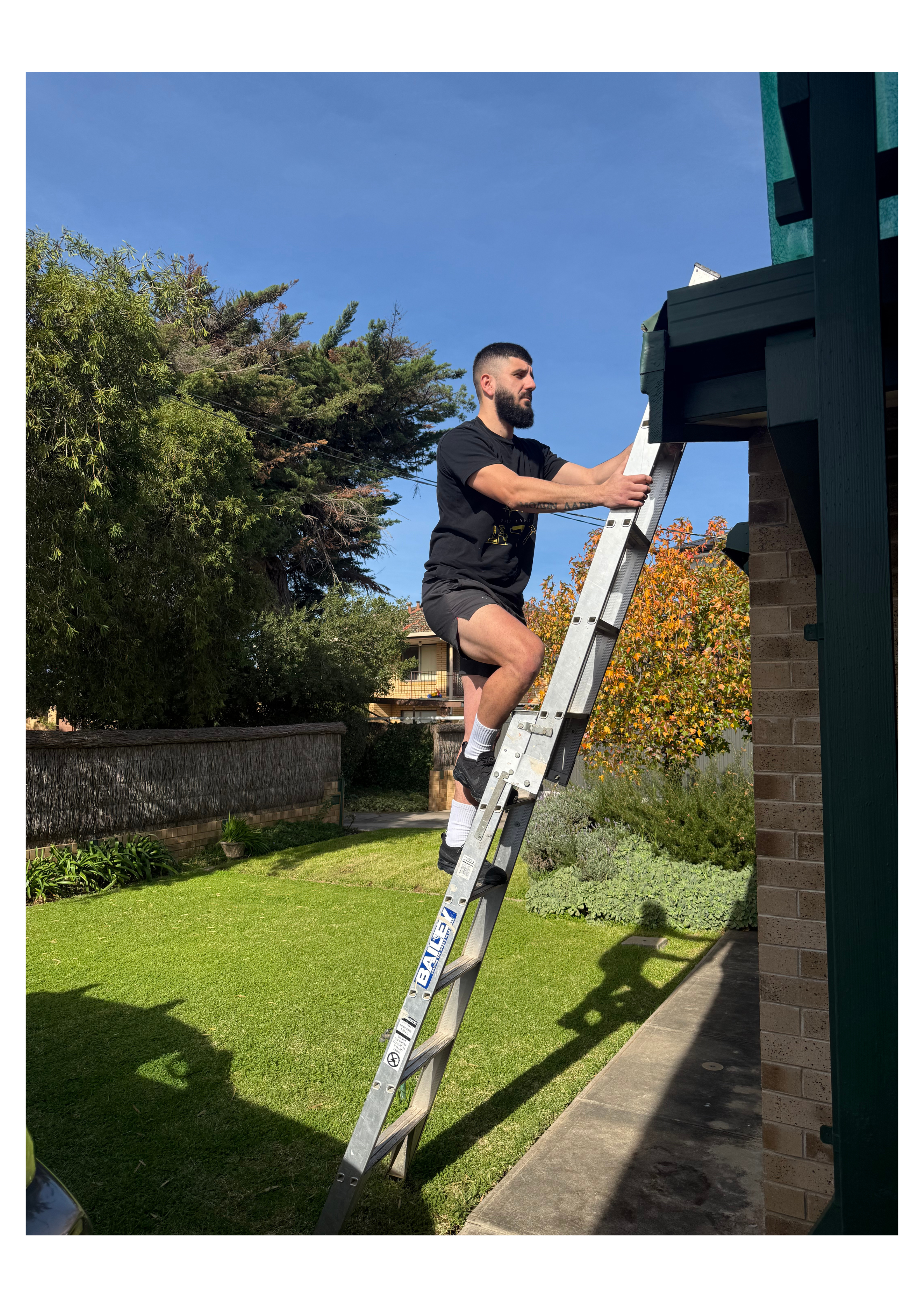 A man with a beard climbing a silver extension ladder against the brick exterior of a house on a lawn under a clear blue sky. Trees and a neighboring house are visible in the background.