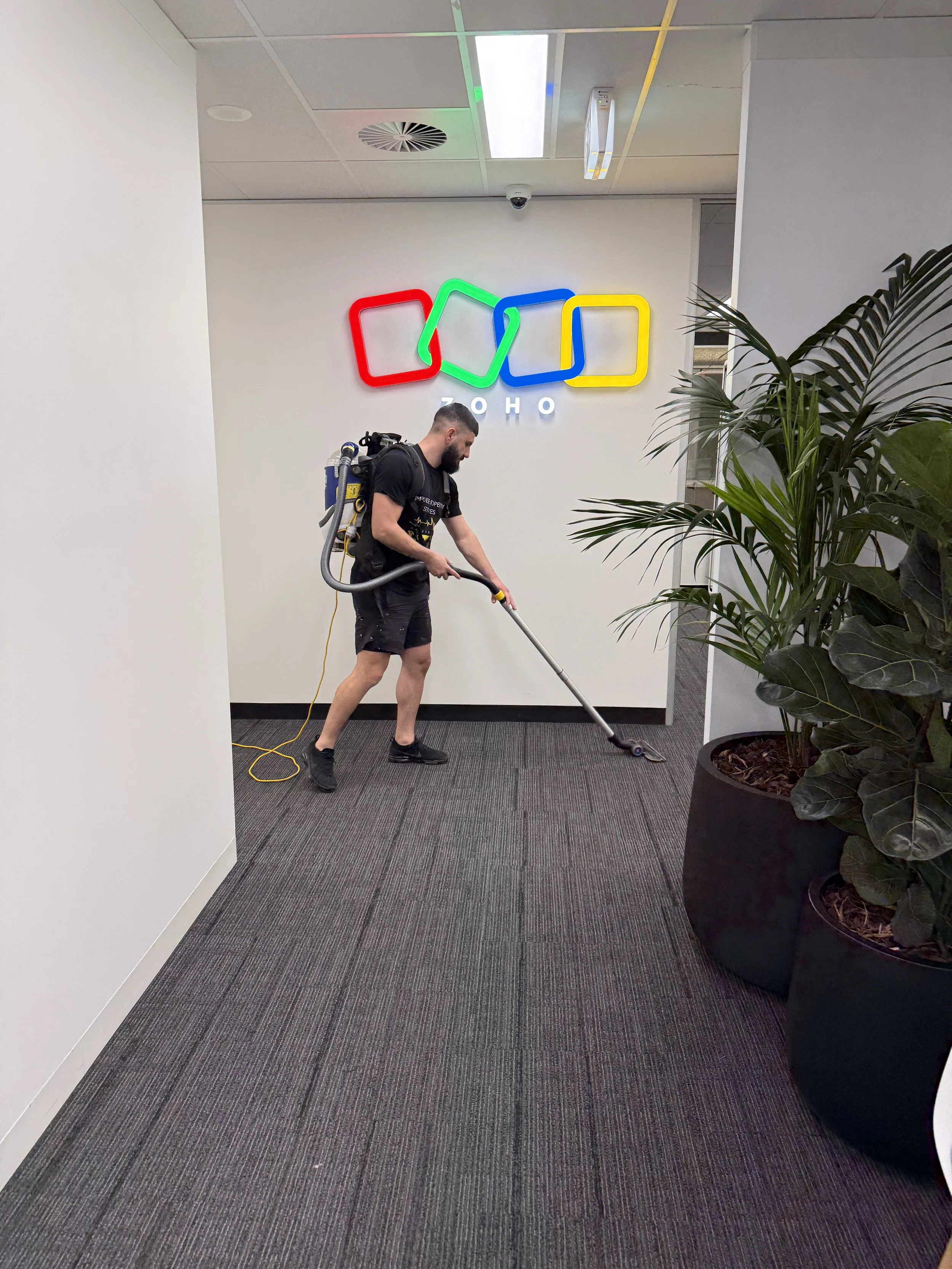 A man with a short beard wearing black shorts and a black t-shirt is vacuuming the carpeted floor of an office hallway. The hallway has white walls, a large potted plant on the right, and a colorful neon sign that reads 'ZOHO' on the wall behind him. The ceiling has fluorescent lights, an air vent, and a security camera.