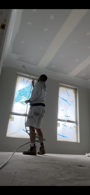 A person painting the ceiling of a room with a roller brush, with windows covered for protection.