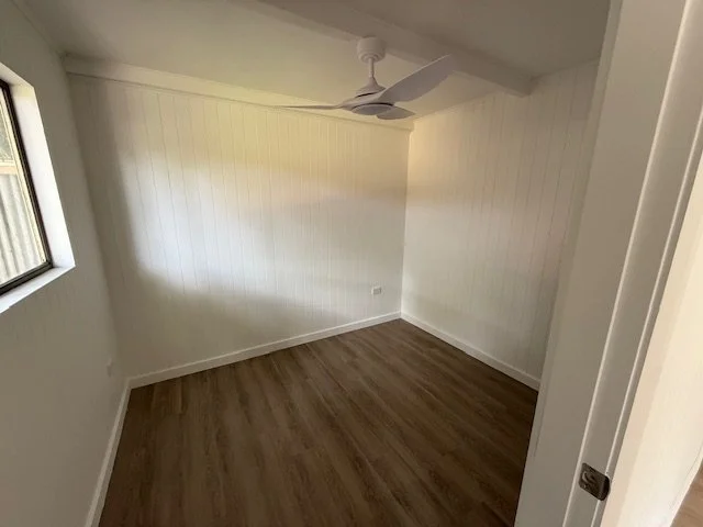 Empty room with white walls, a white ceiling fan, wood laminate flooring, a window on the left, and a partially open door on the right.
