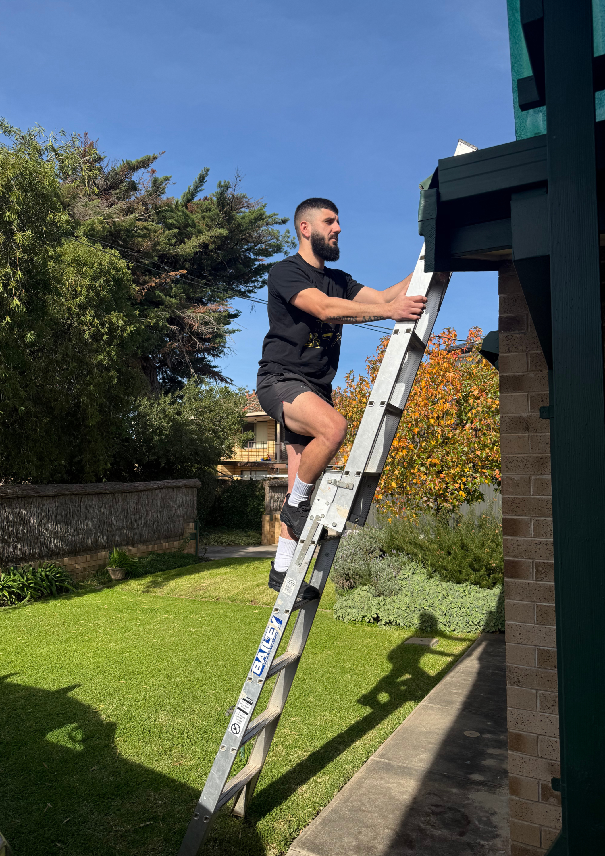 A man with a beard and tattoos wearing black shorts and a black T-shirt is standing on a ladder, painting the exterior of a brick building in a backyard.