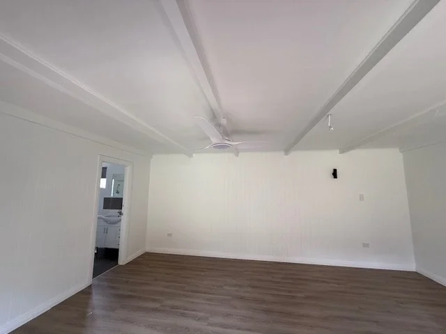 Empty room with white walls, wood flooring, ceiling fan, and an open doorway leading to a small room with a sink and window.