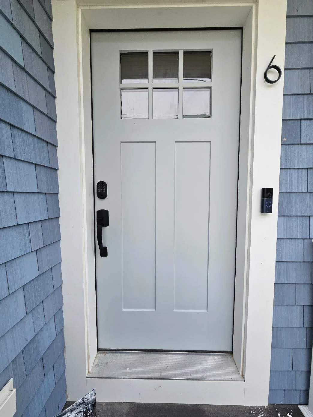 White front door with a small window at the top, black handle, and keypad lock, surrounded by blue siding and white trim, with house number 6 on the right.