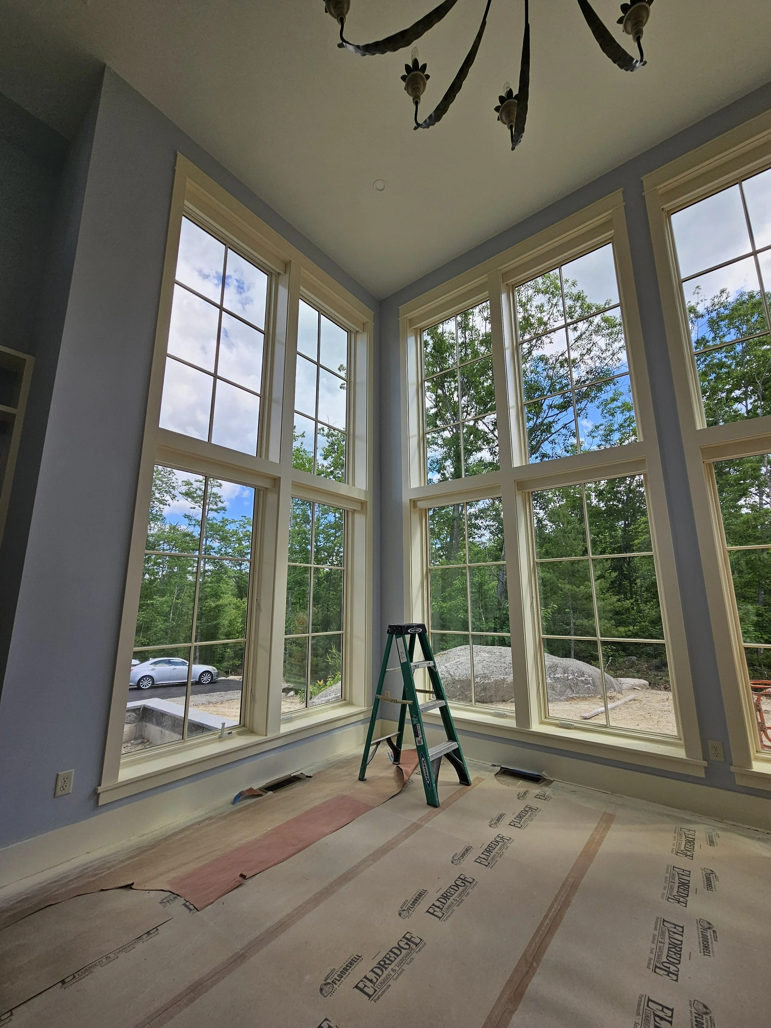 Interior view of a room under construction with large floor-to-ceiling windows, a ladder in the center, and construction materials on the floor.