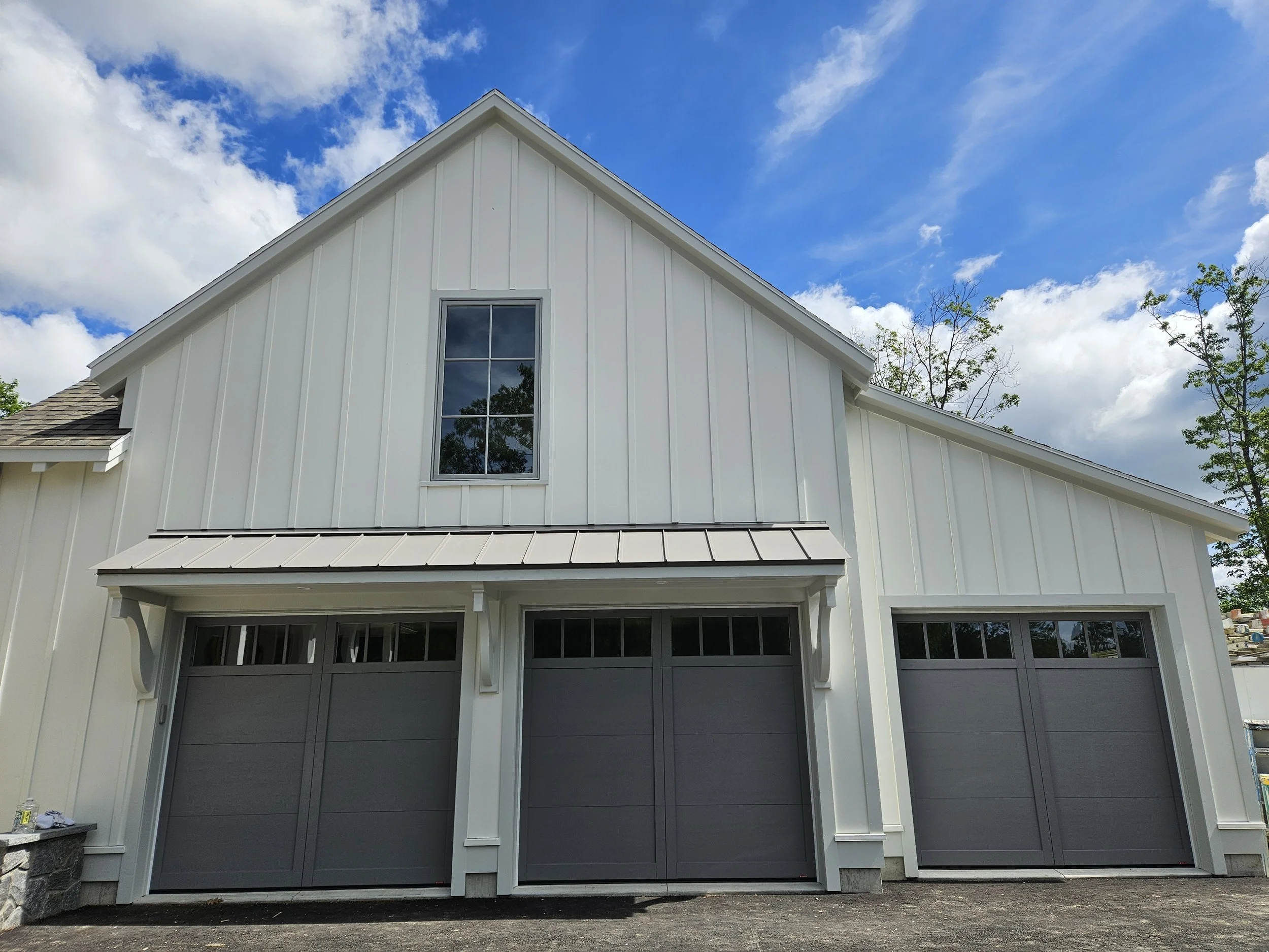 The image shows a two-story white farmhouse with a steep triangular roof, a large window on the second floor, and a garage door on the first floor. There are some trees and a partly cloudy sky in the background.