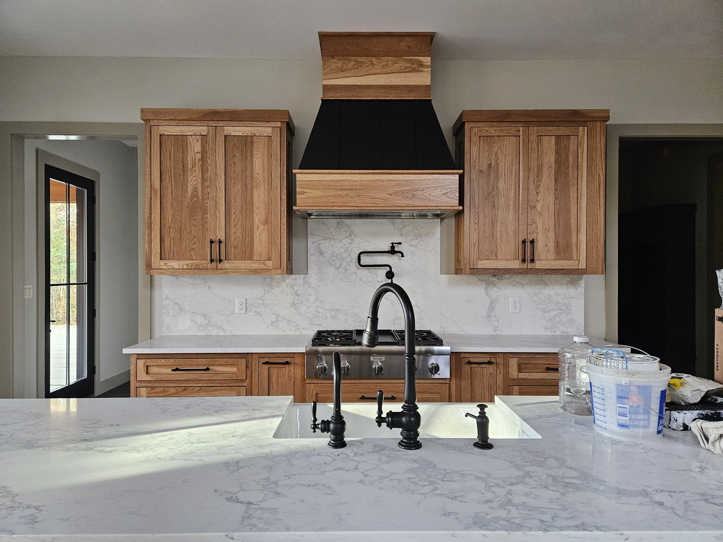 Kitchen with wooden cabinets, marble countertop, black faucet, and stove. Window on the left side, and some cleaning supplies and water bottles on the counter.