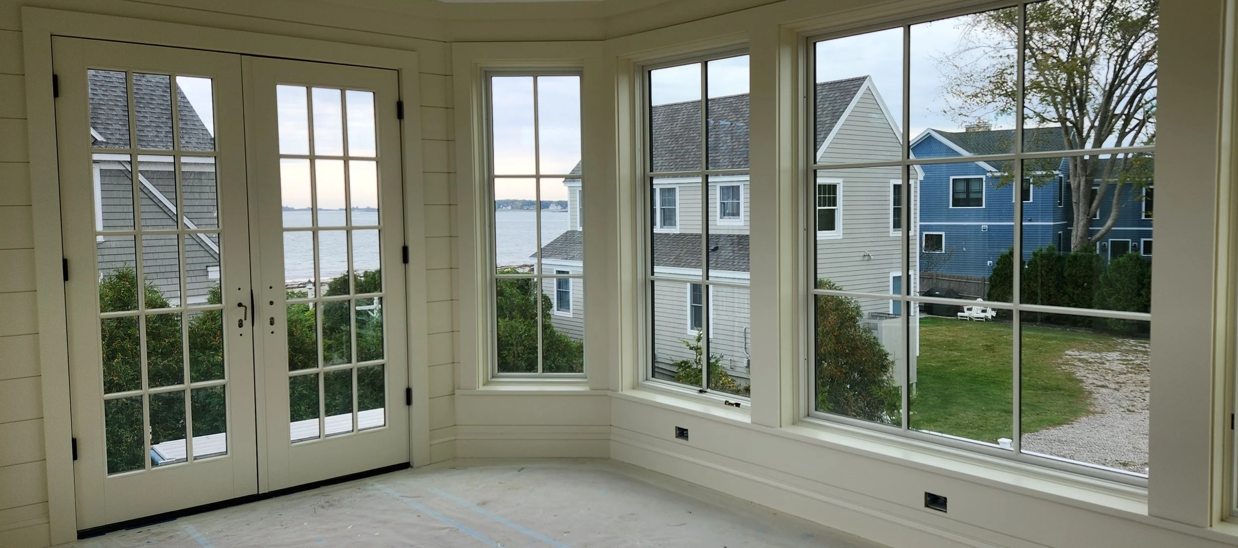Interior of a house with a large window and glass door, overlooking neighboring houses and a body of water outside.