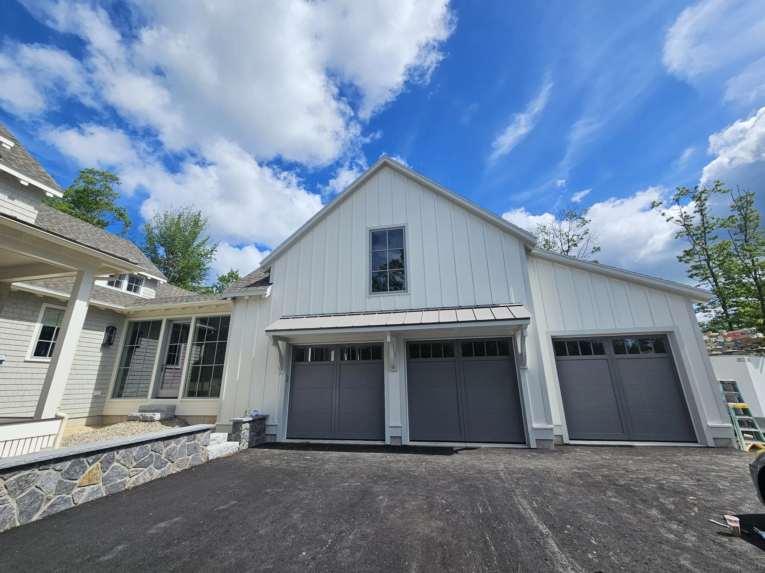 A residential house under construction with a white exterior and large windows, an attached garage with closed doors, a stone border in front, and trees surrounding the property. The sky above is partly cloudy with bright blue skies.