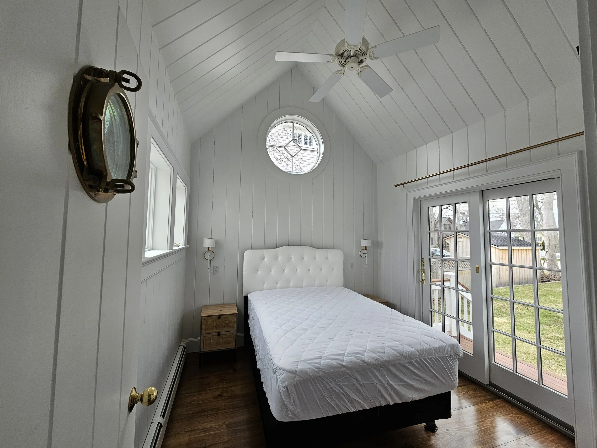 A bedroom with white paneled walls and ceiling, a ceiling fan, a window with an arched top, a bed with a white padded headboard and mattress, two nightstands with lamps, and a sliding glass door leading to an outdoor space.