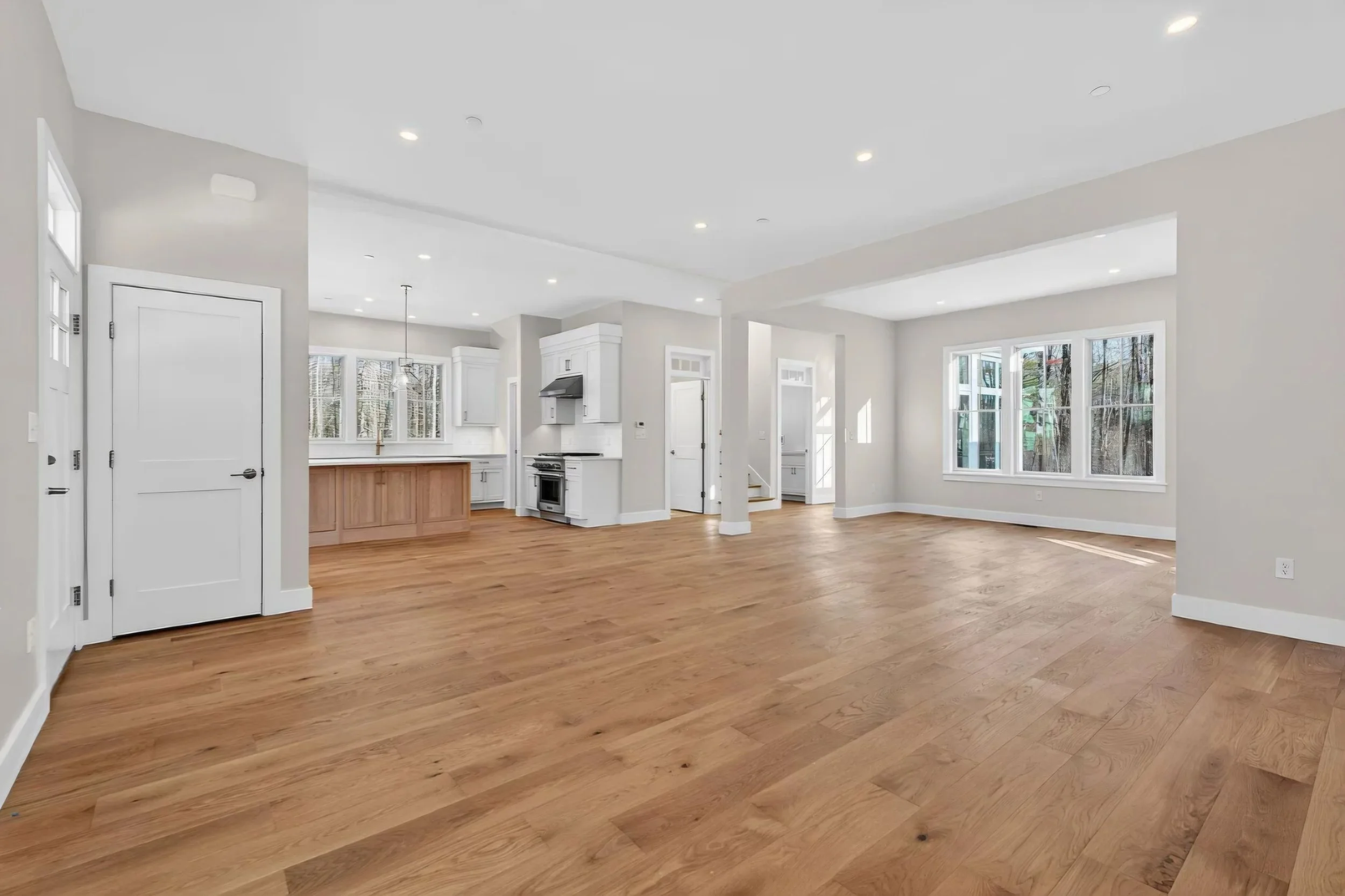 Open floor plan living room and kitchen with large windows, hardwood flooring, white walls, and modern appliances.