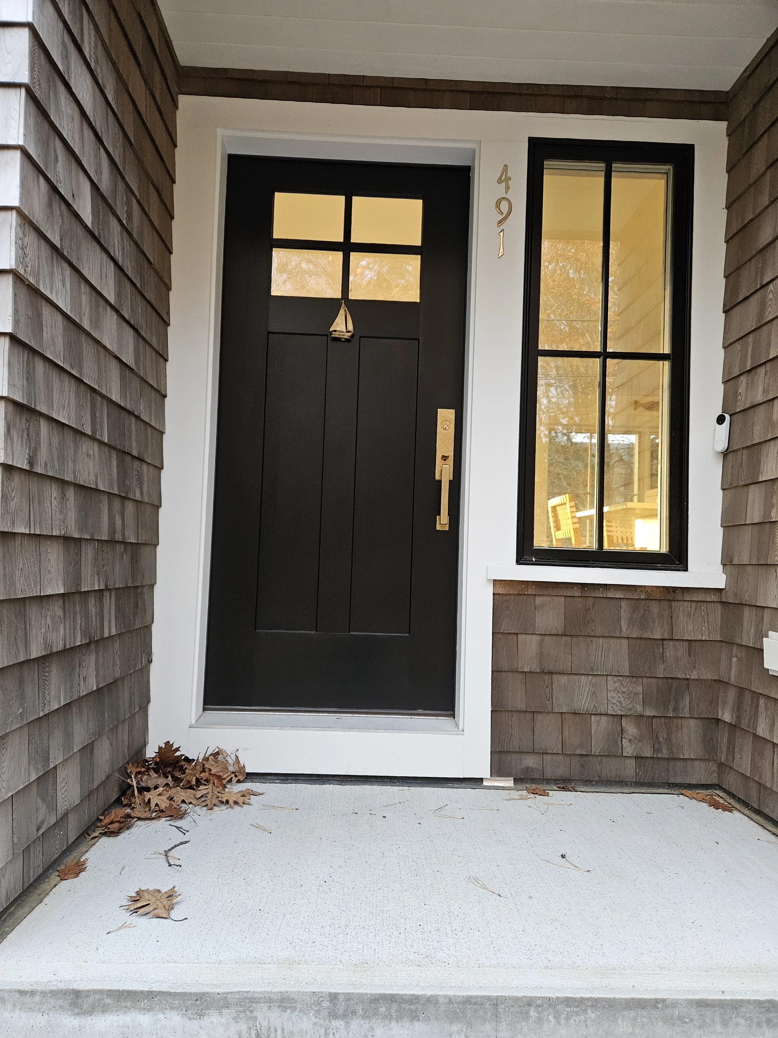 Front porch of a house with a black door, a window with black frame, and house number 491. The porch has a light-colored concrete floor with some fallen leaves.