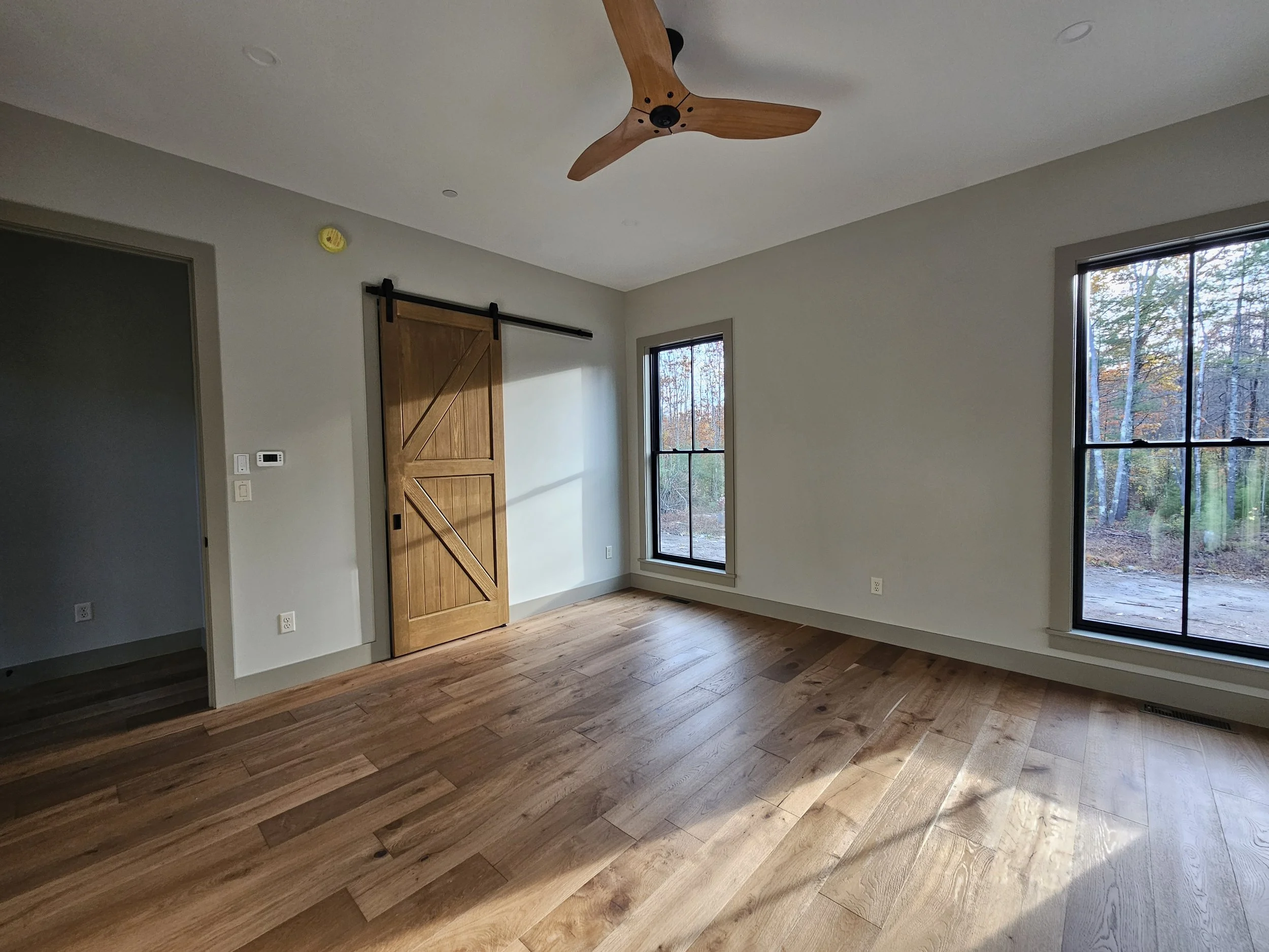 Empty room with wooden floors, two large windows showing trees outside, a sliding barn door, and a ceiling fan with wooden blades.